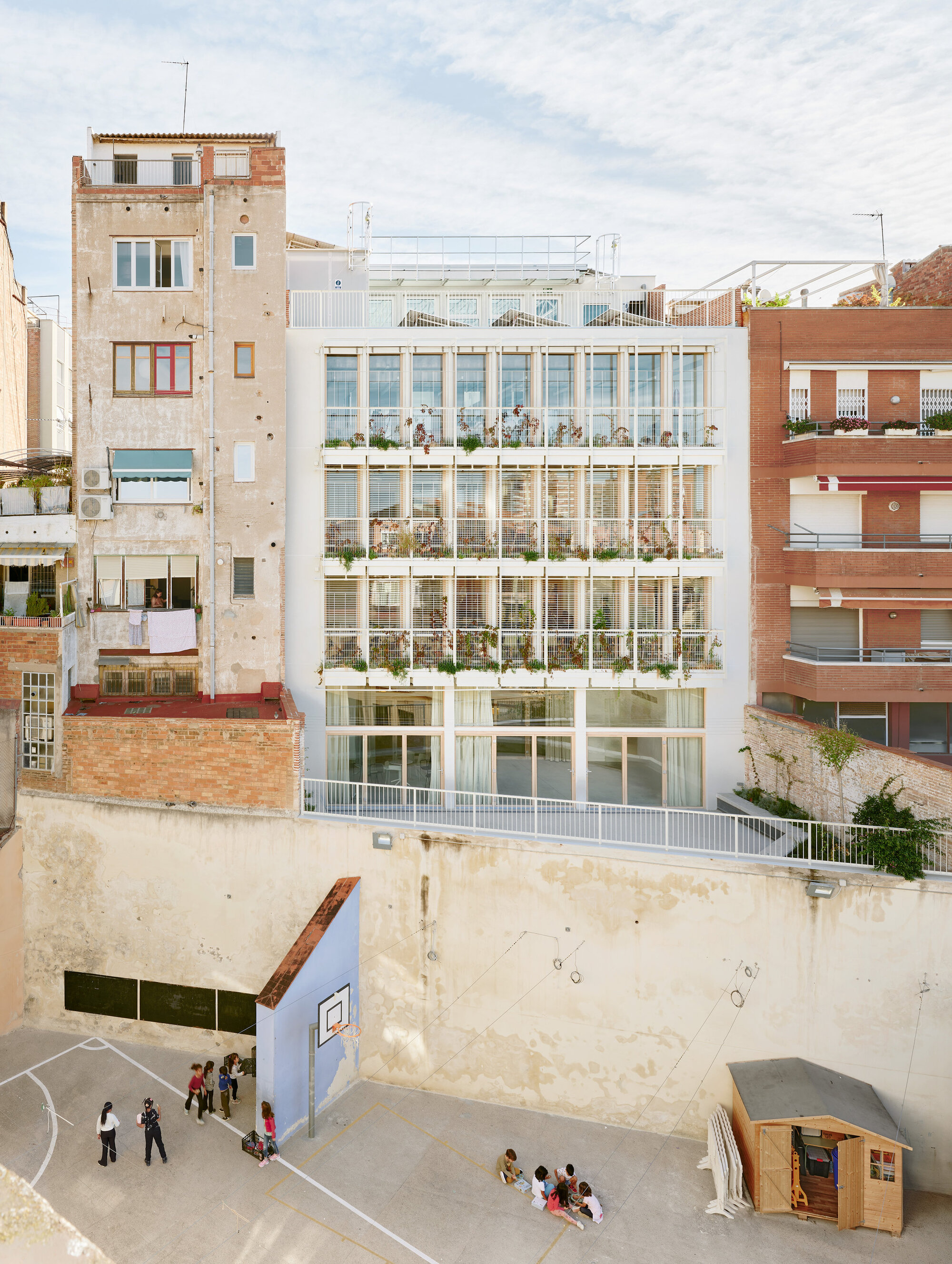 White gridded facade with planted balconies rising above a schoolyard wall, with children playing near a basketball hoop in afternoon sun
