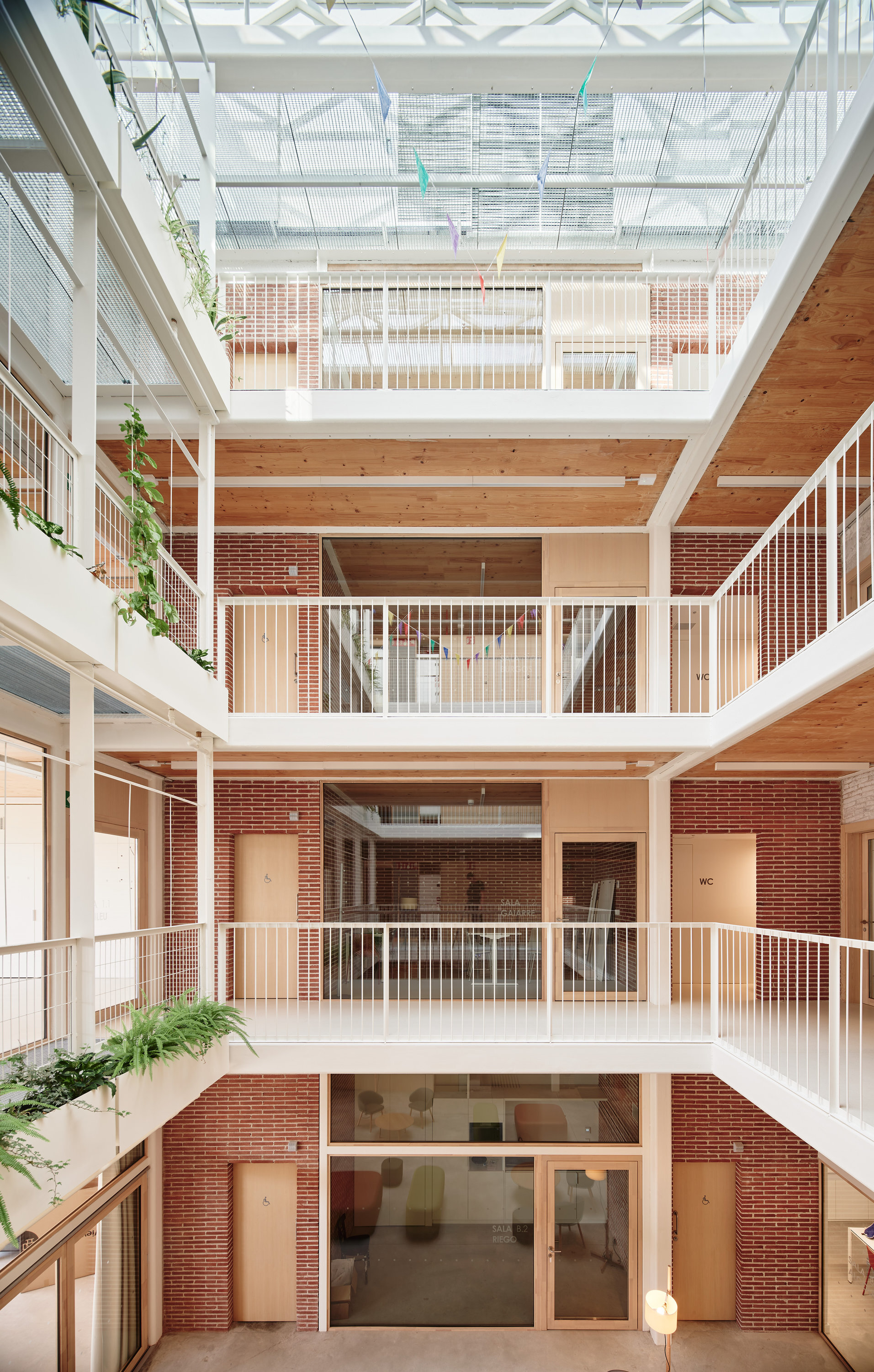 Interior atrium with timber ceilings, red brick walls and white walkway railings rising toward a glazed skylight hung with paper bunting