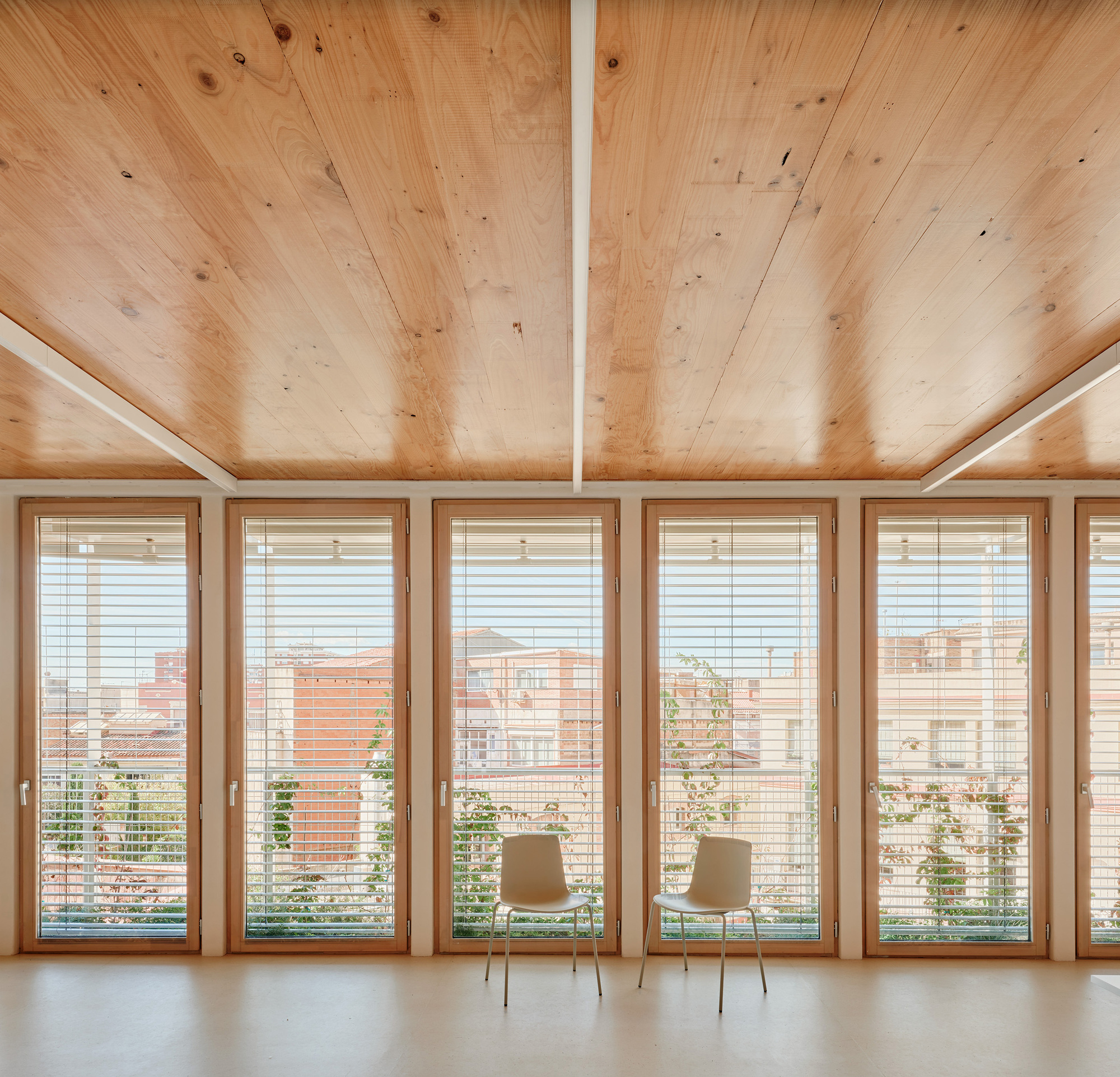 Timber-framed tall windows with slatted blinds and two white chairs beneath a pine plank ceiling, vines visible outside in daylight
