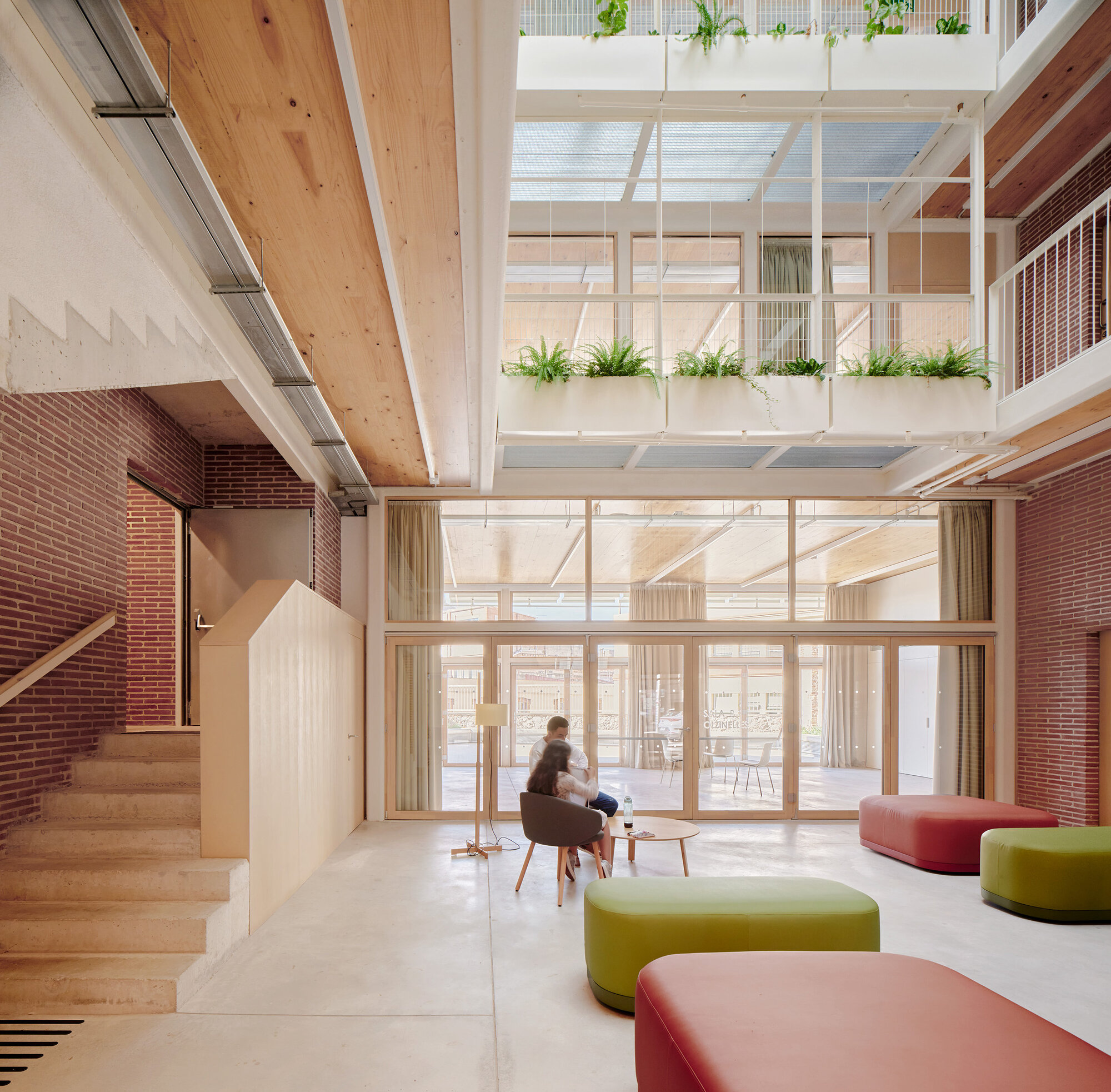 Double-height lobby with brick walls, a timber stair and green and red ottomans, with two people seated near planters overhead