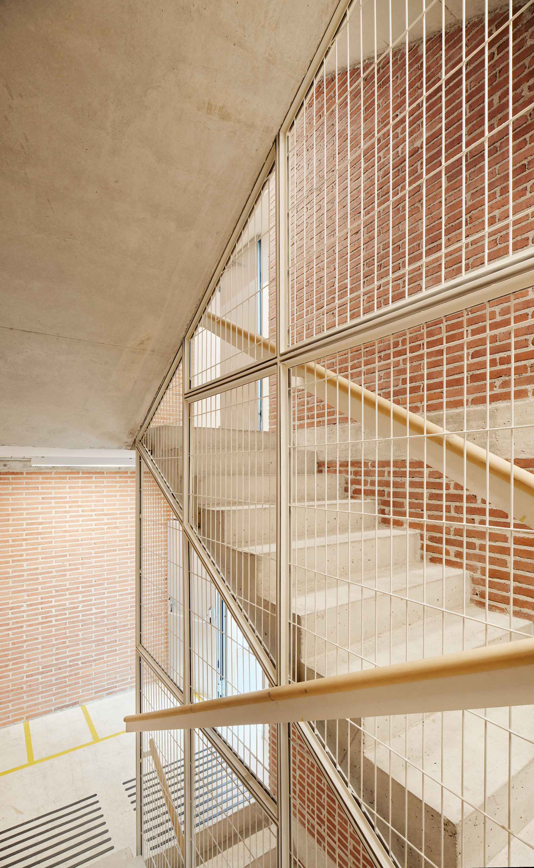 Concrete stair behind a white steel mesh screen with a timber handrail, set against a red brick wall