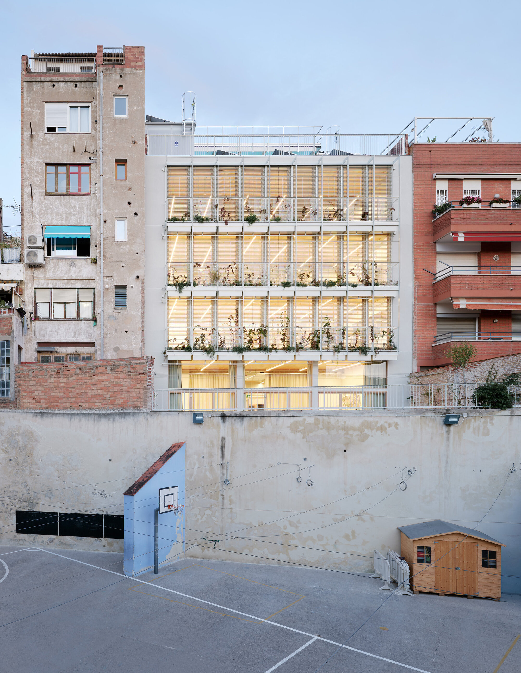 Evening view of the lit, plant-lined glass facade rising above a schoolyard basketball hoop, wooden shed and retaining wall