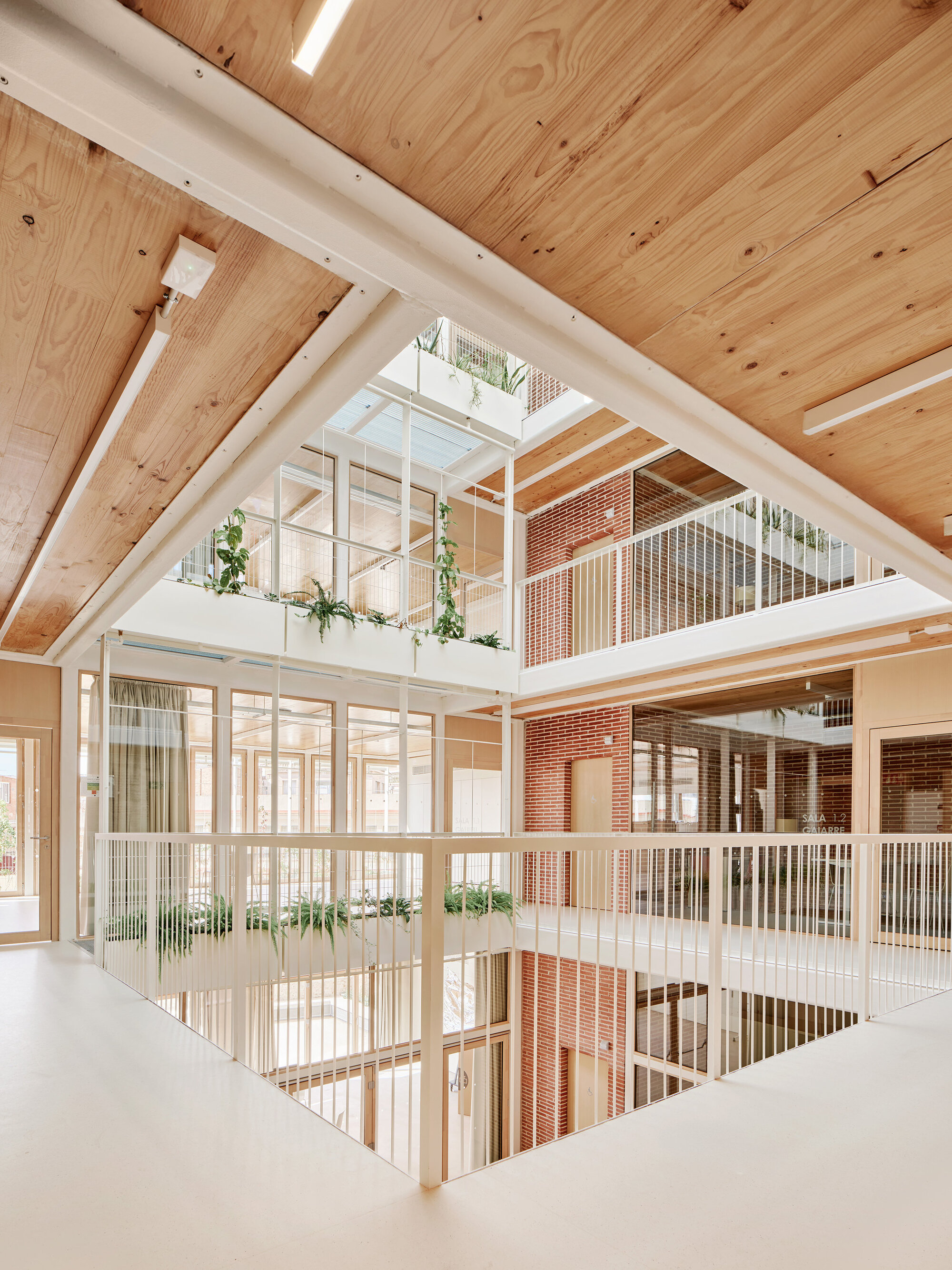 View from a walkway across a white balustrade into the atrium, with a plywood ceiling, brick walls and hanging plants