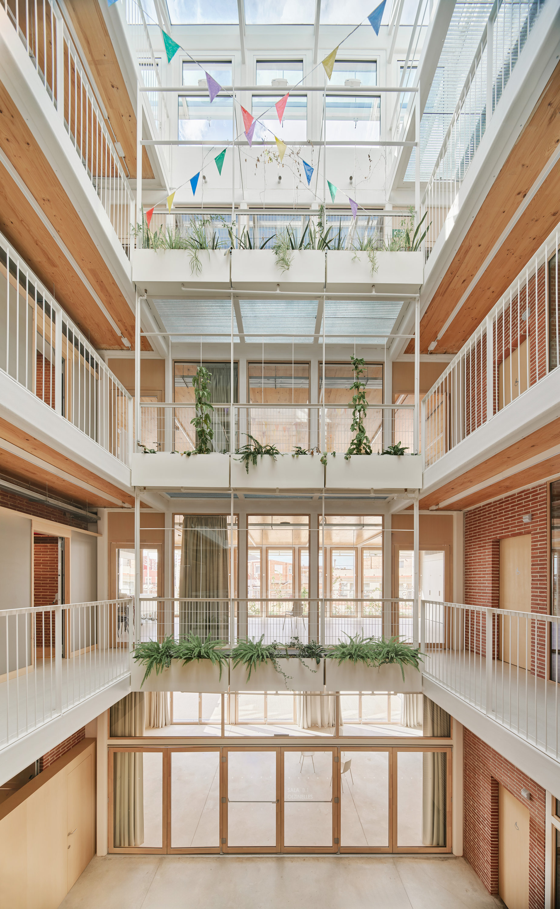 Central atrium seen from the floor, with planter-lined galleries, timber soffits and brick walls rising to a skylight hung with colored bunting