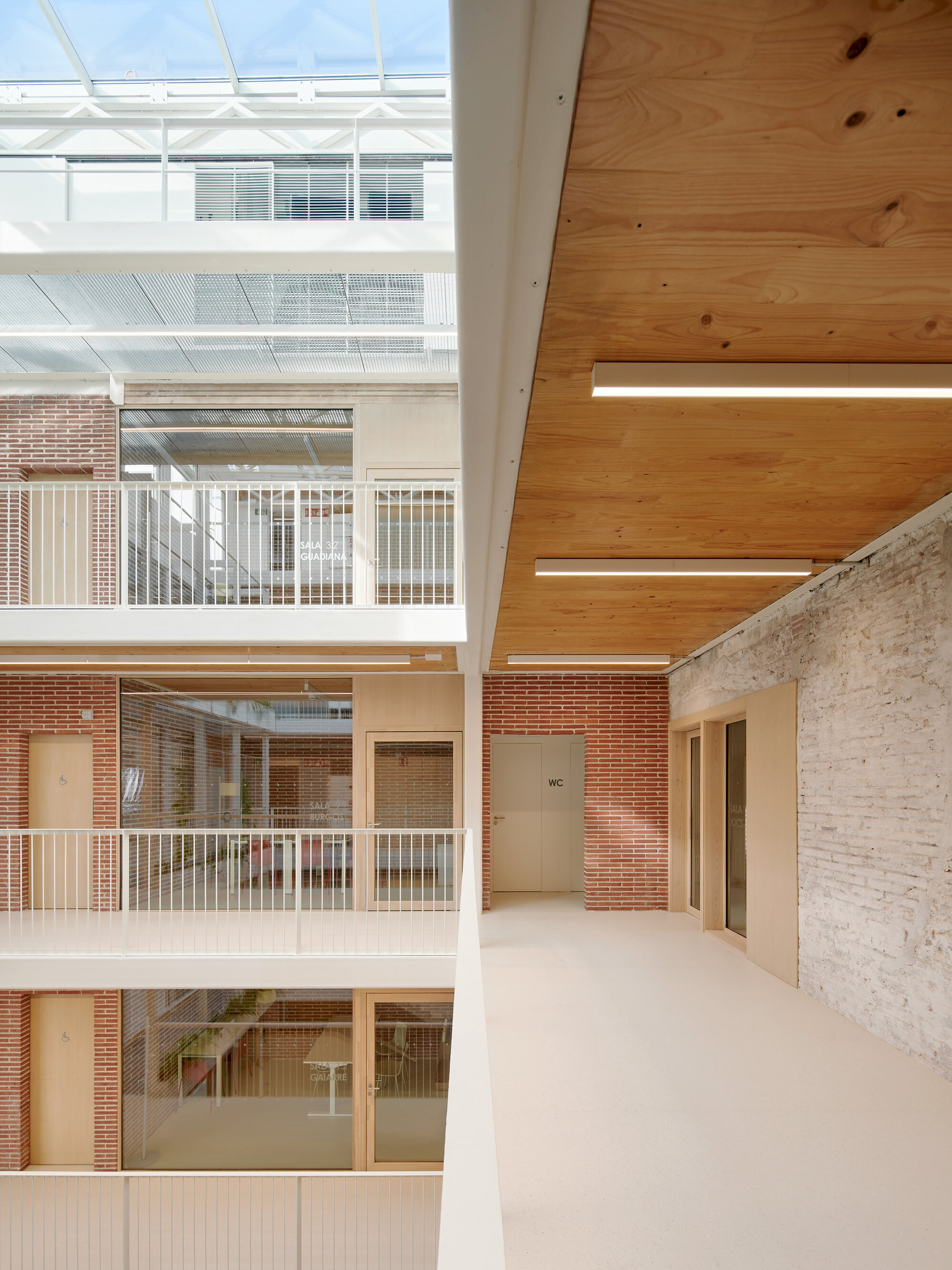 Upper gallery with plywood ceiling and linear light fixtures beside red brick walls, white railings and a glazed roof overhead in daylight