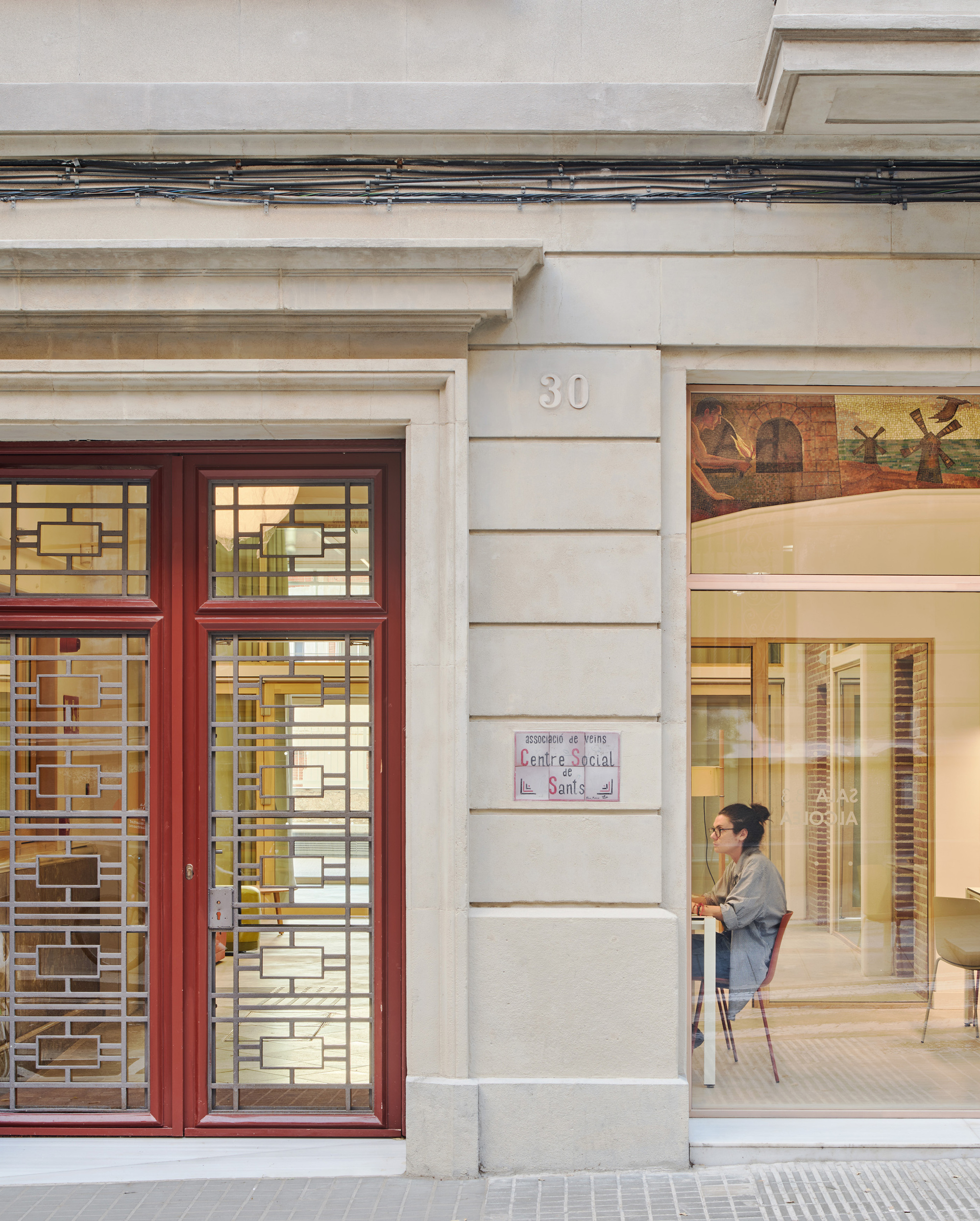 Street facade with a red-framed door, patterned metal grille and a woman seated at a table behind the glazed opening