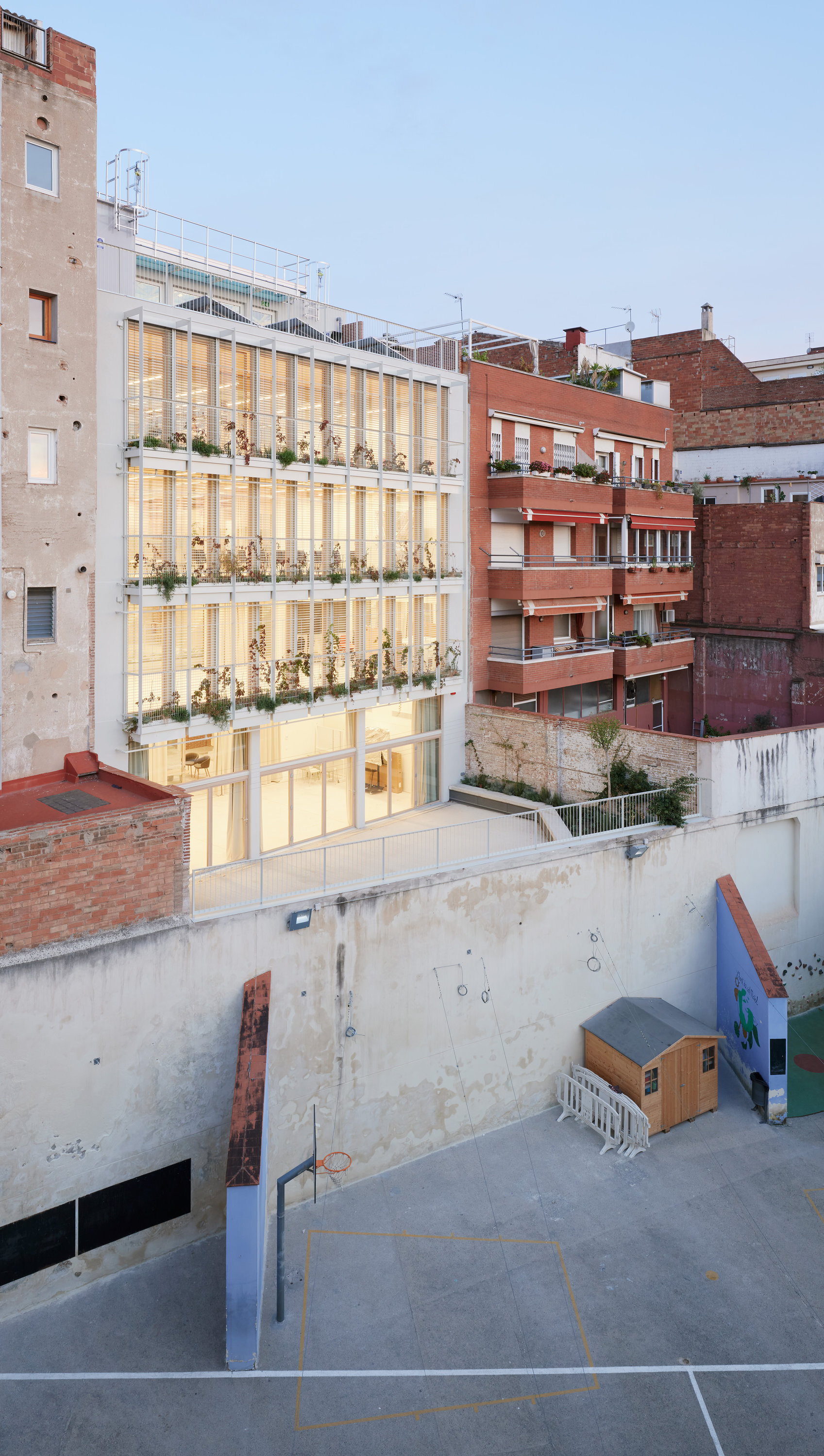 Rear facade of glazed tiers with planted balconies, seen over a courtyard wall at dusk with a small timber shed