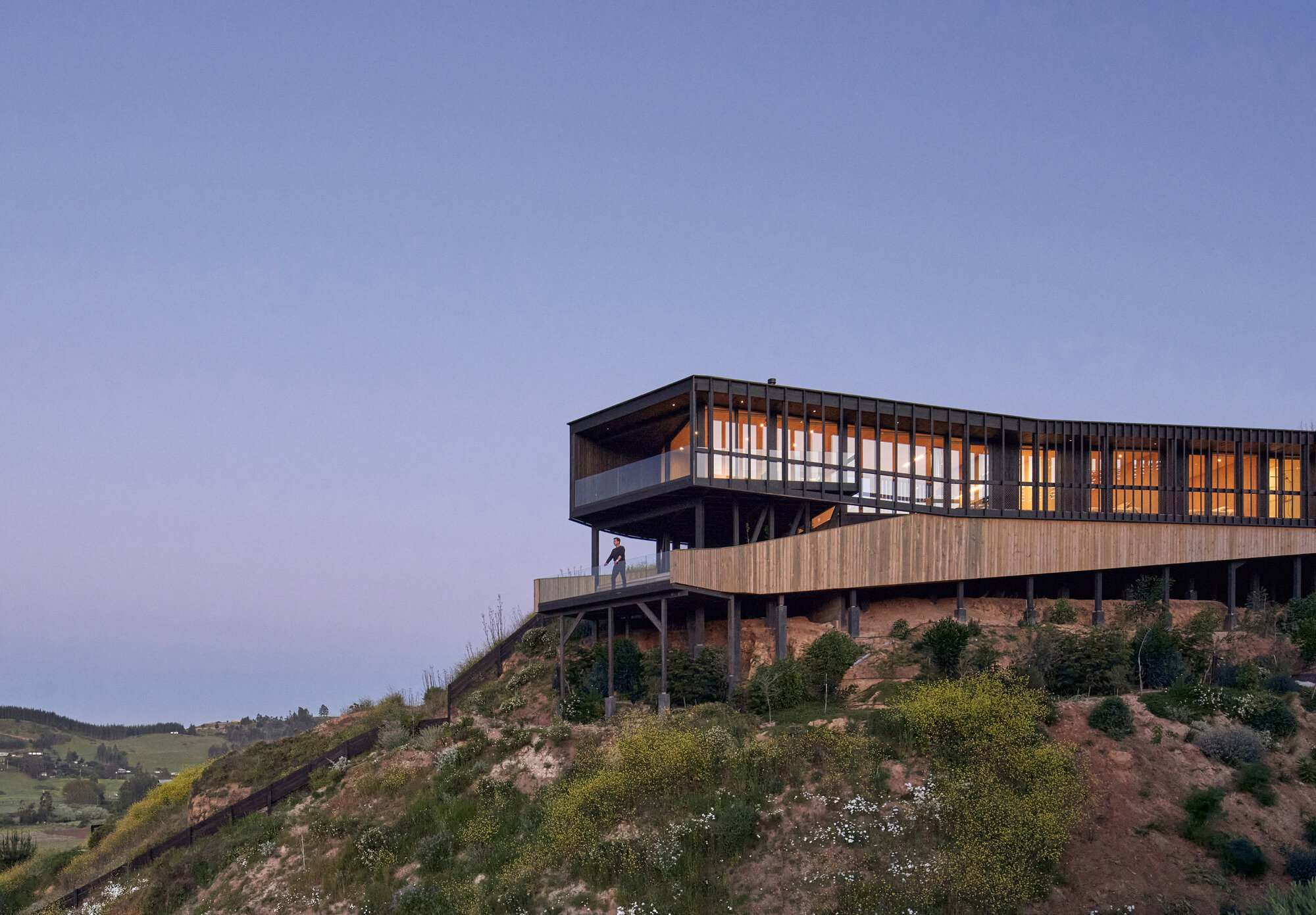 Cantilevered timber house on stilts above a scrubby clay slope, windows glowing amber under a violet dusk sky