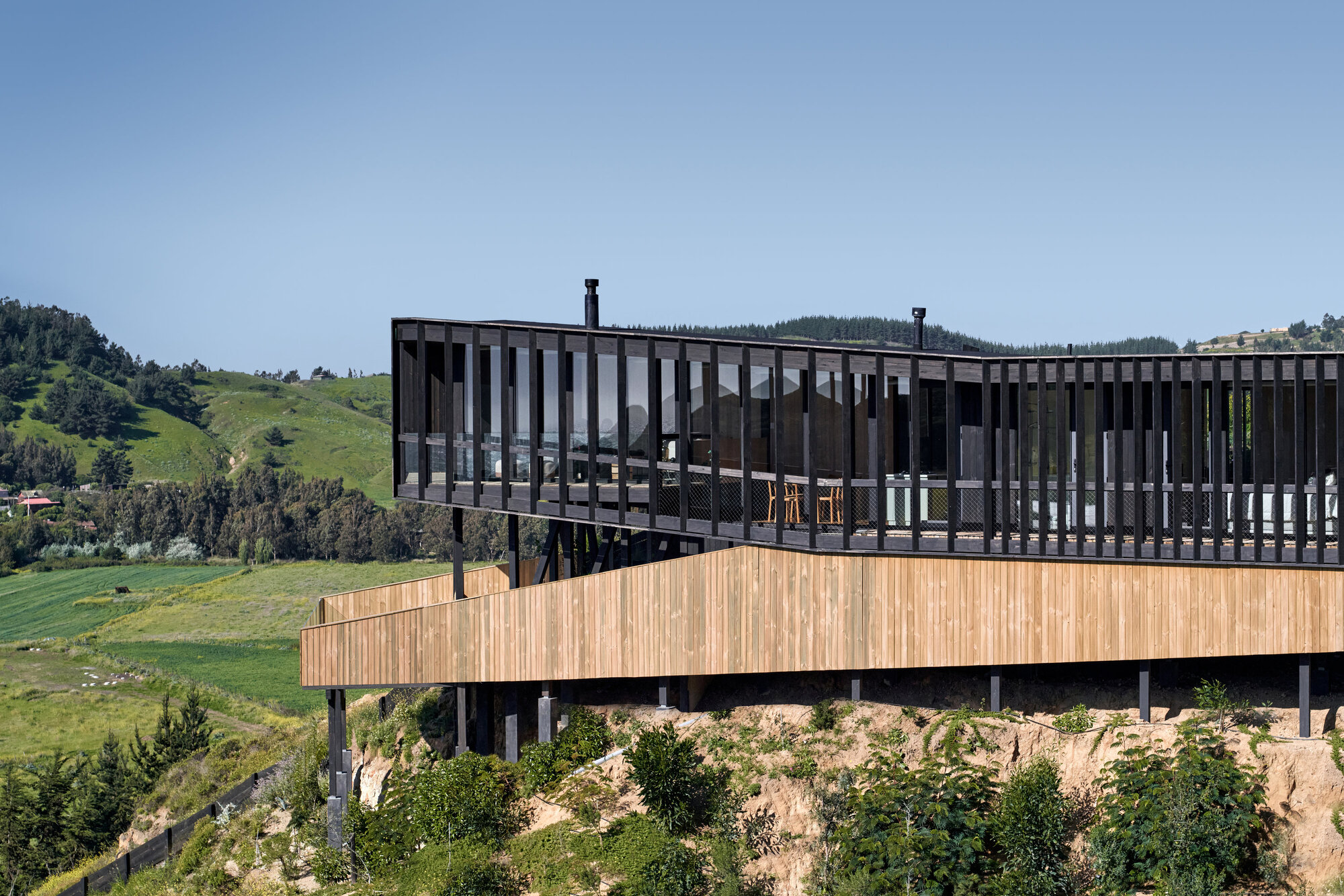 Side view of the slatted dark timber upper volume above a pale wood-clad underside, with green hills beyond in daylight