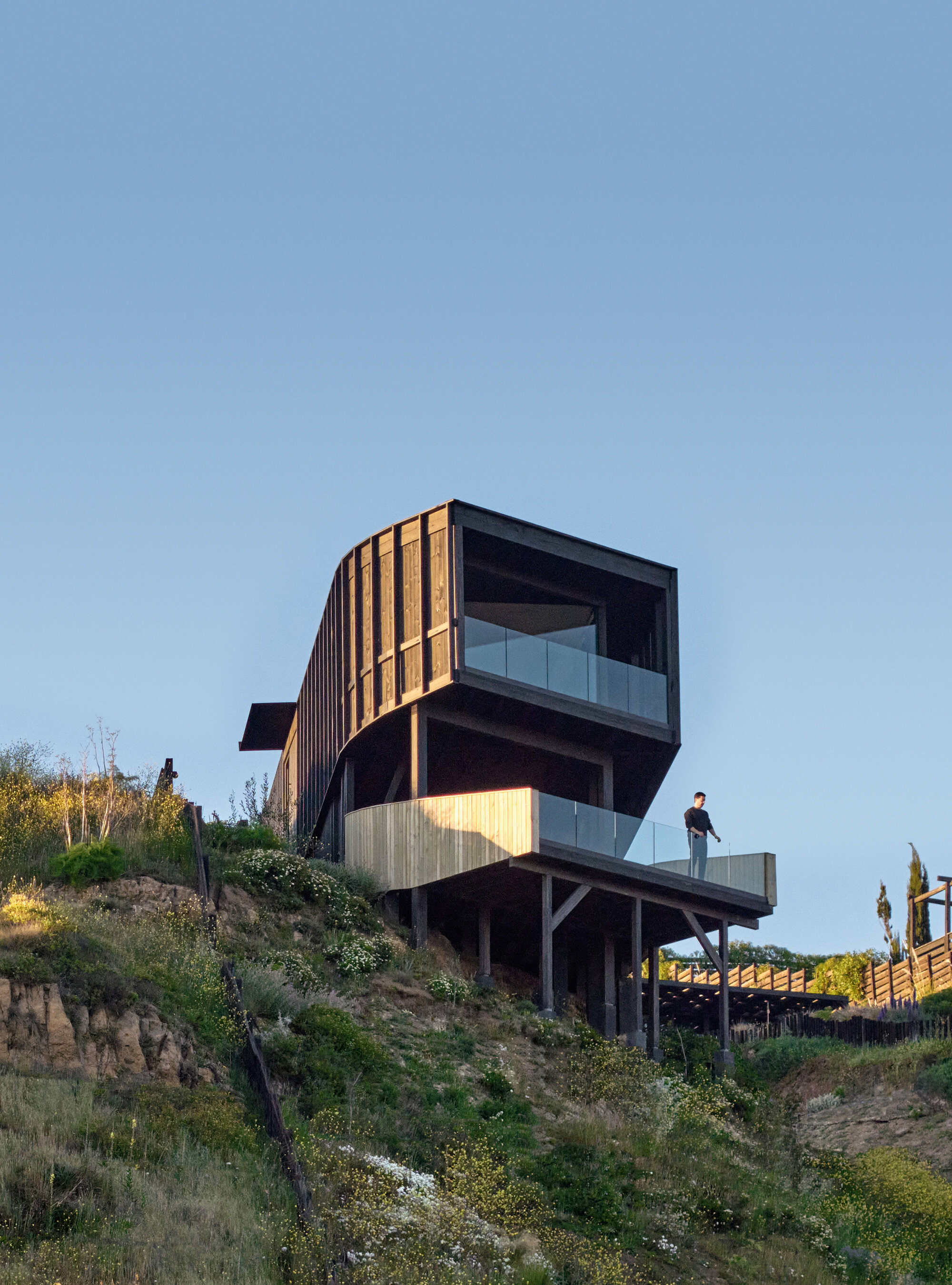 Low angle view of the stepped timber volume with glass balustrade and a person standing on the deck at sunset