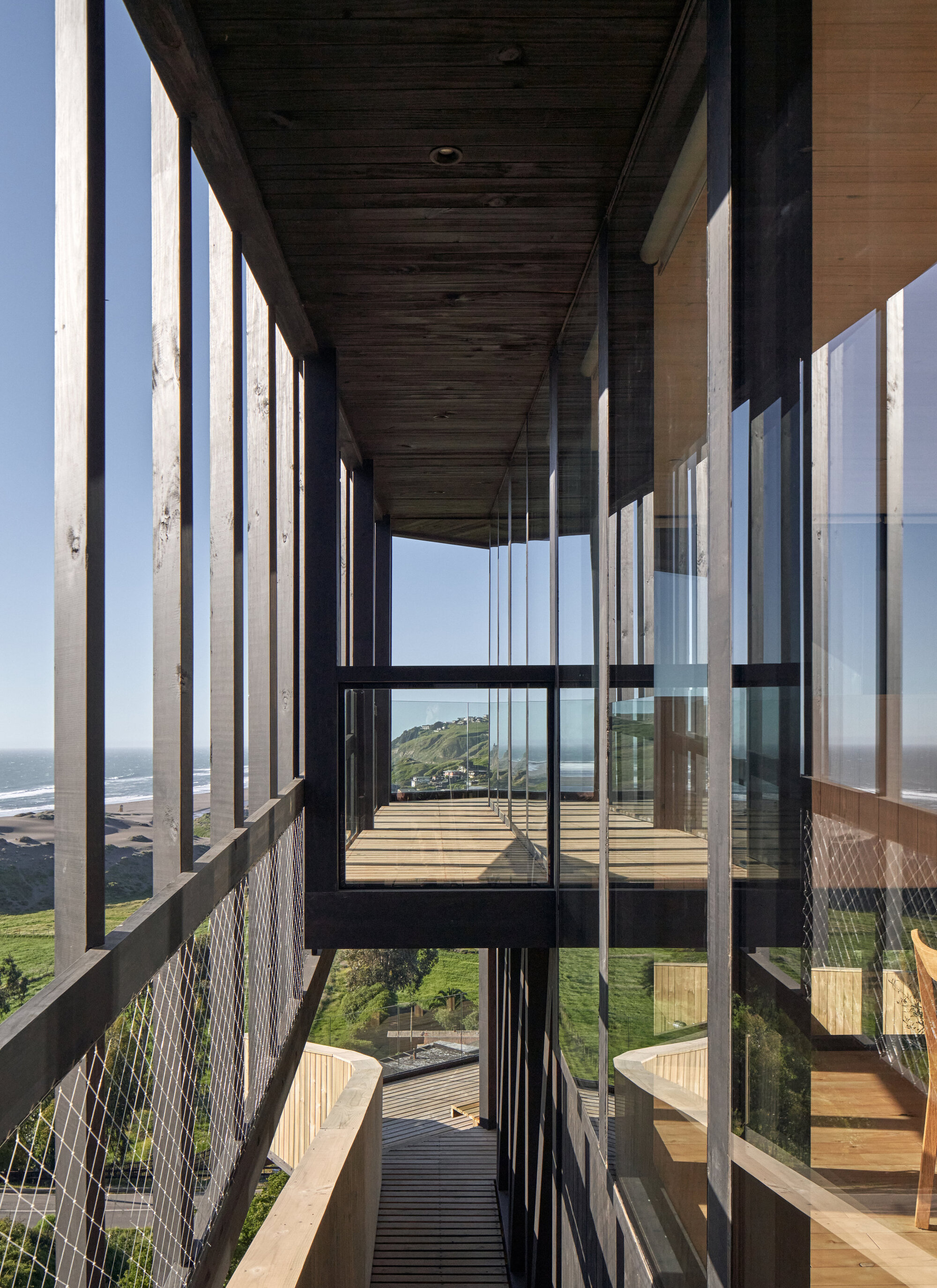 Narrow timber gallery with vertical slats, cable mesh guard and a glimpse of the ocean and beach beyond