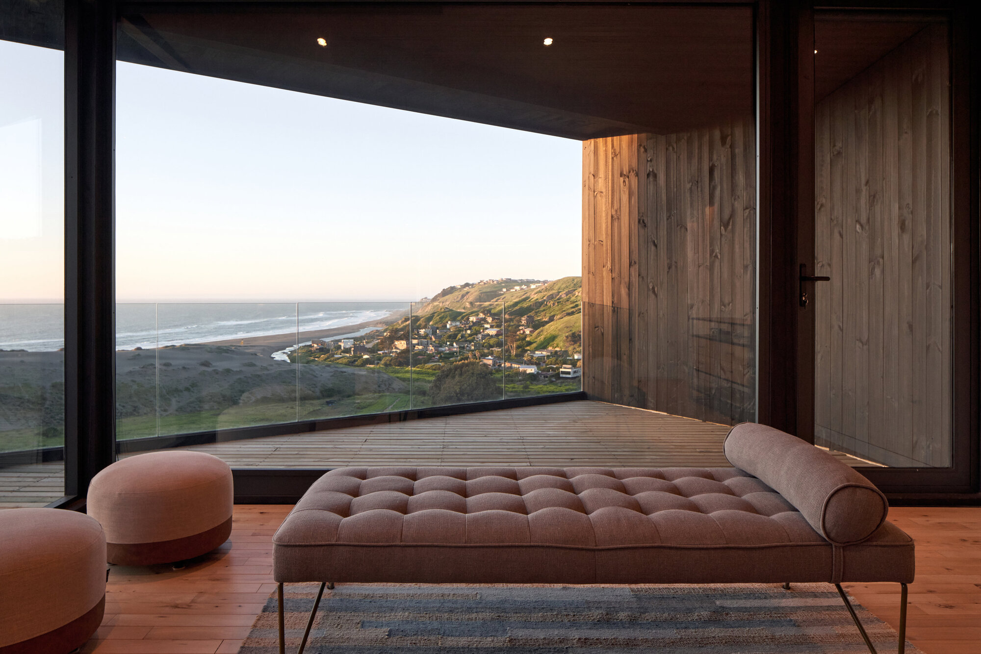 Living room corner with tufted daybed and round pouf facing a glass wall over the coastline at dusk