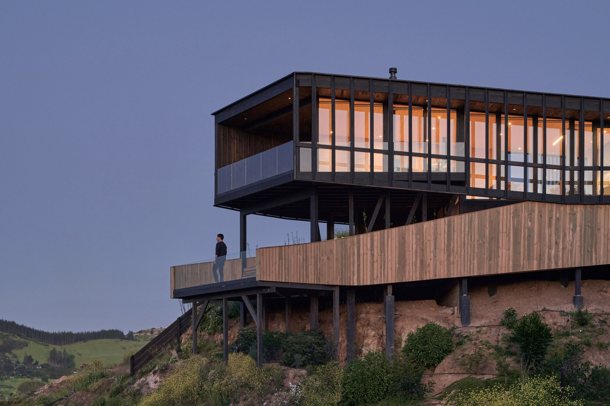 Close view of the elevated timber house on steel and timber posts, a person standing on the deck at blue hour
