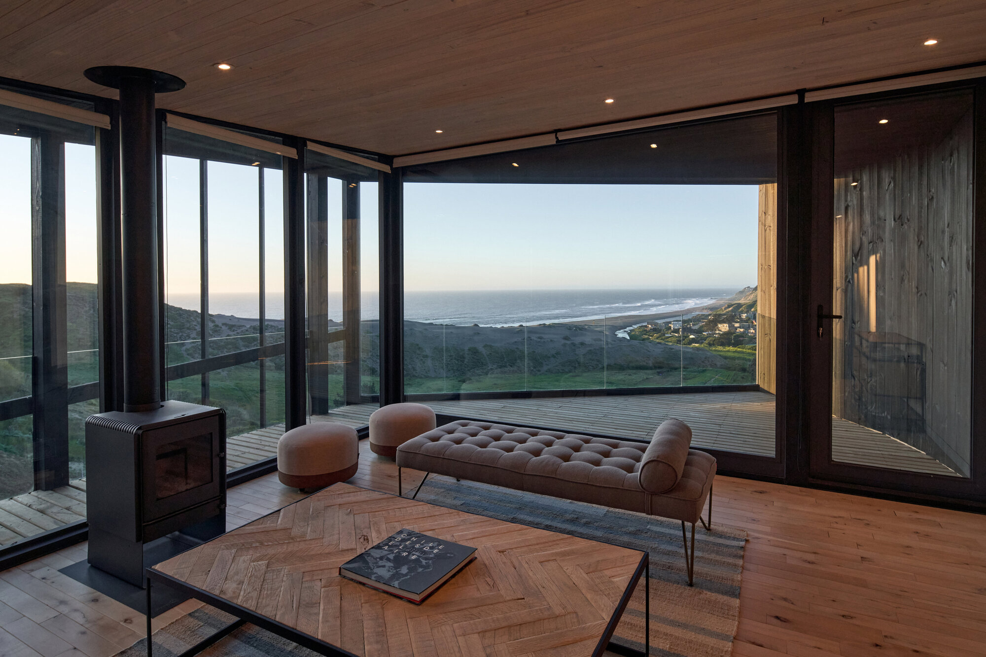 Living area with a black wood stove, tufted bench and herringbone coffee table facing a glazed corner and the ocean at dusk