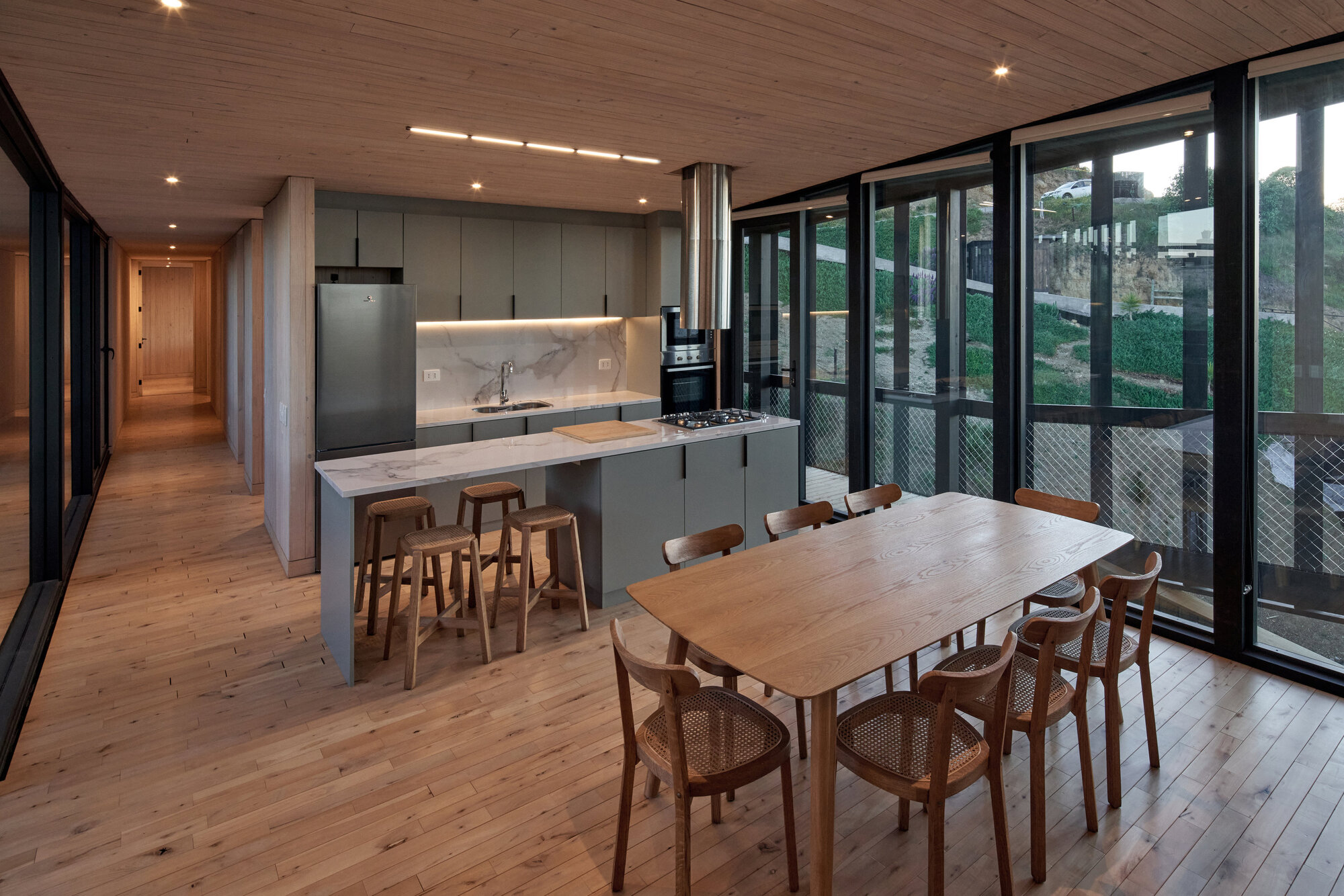 Kitchen with marble-topped island, wooden stools and dining table beside floor-to-ceiling glazing and a timber plank floor