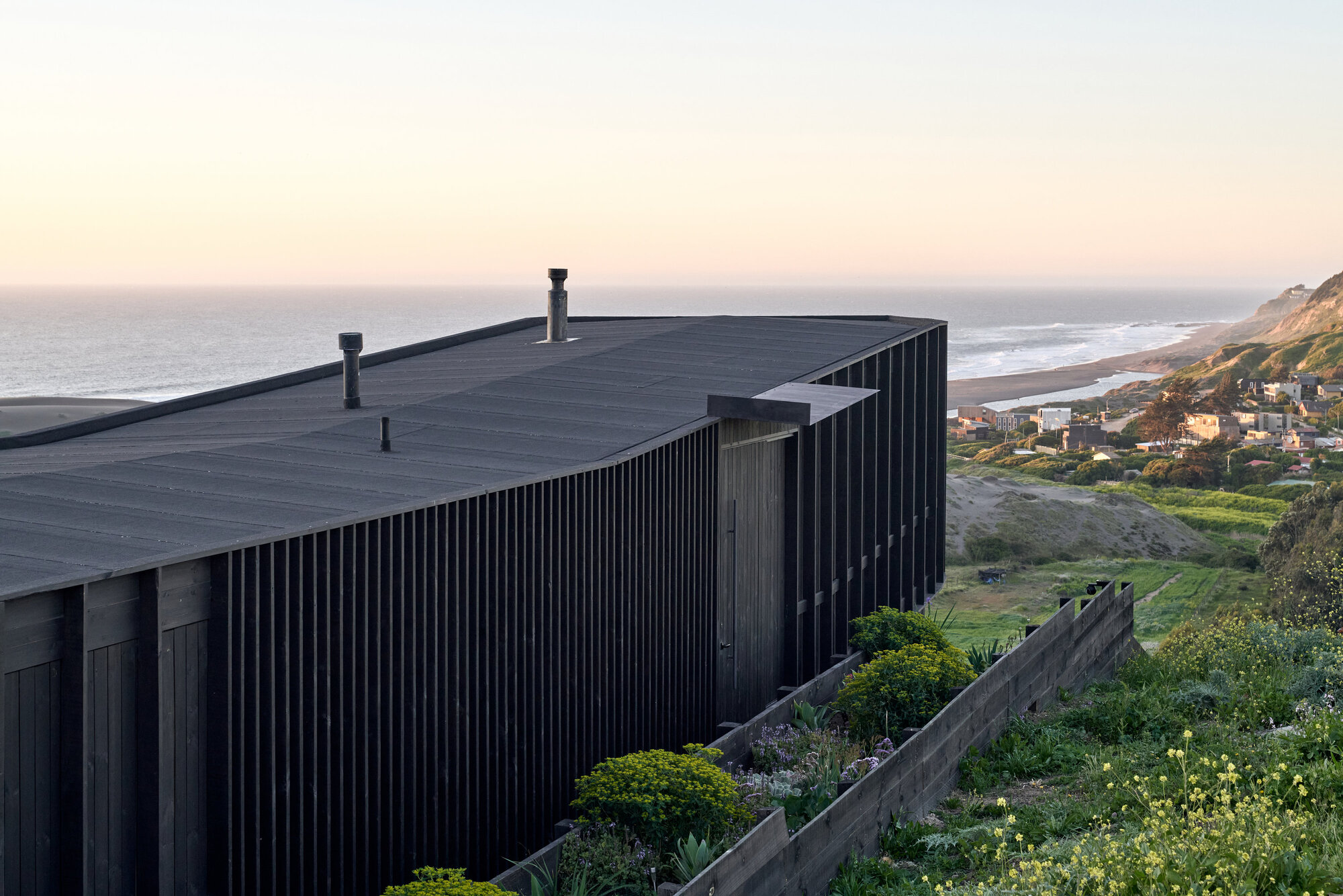 Dark charred timber cladding and flat roof with two flues, seen above planted terraces and the sea at sunset