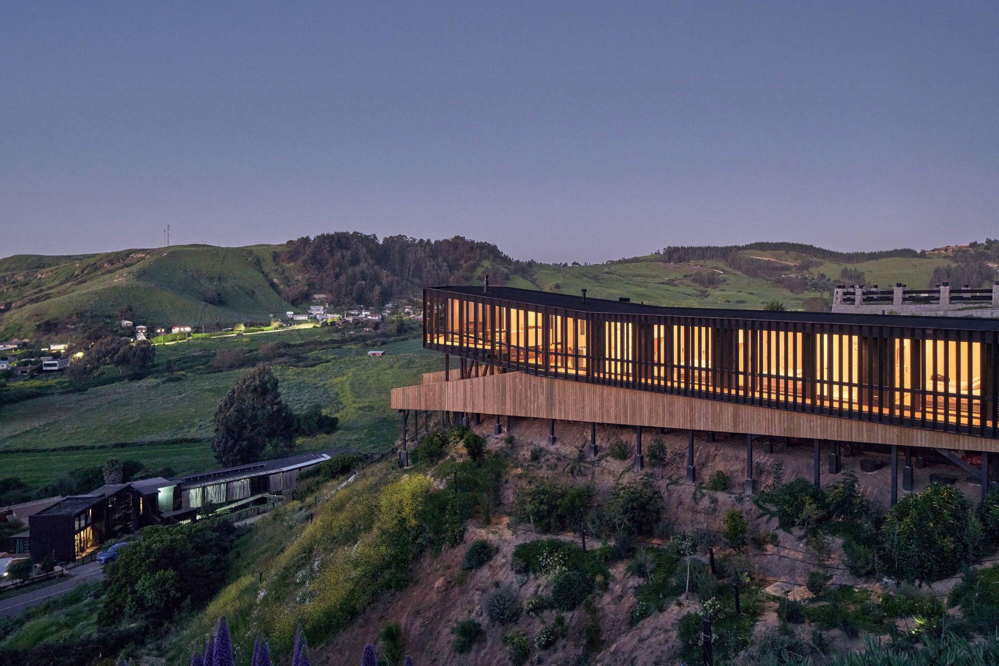 Long glowing timber-clad volume raised on slender steel posts above a steep planted slope at blue hour