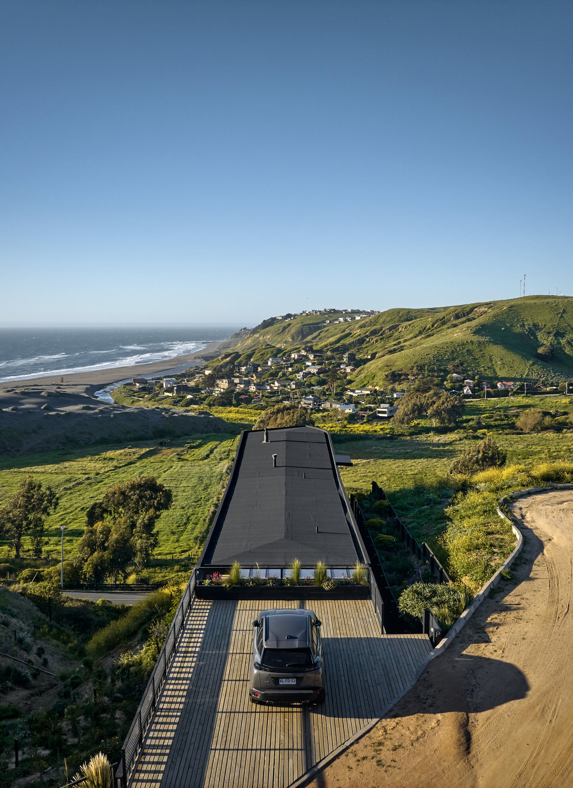 Aerial view of the black flat roof and timber deck with a parked car, green hills and coastline behind