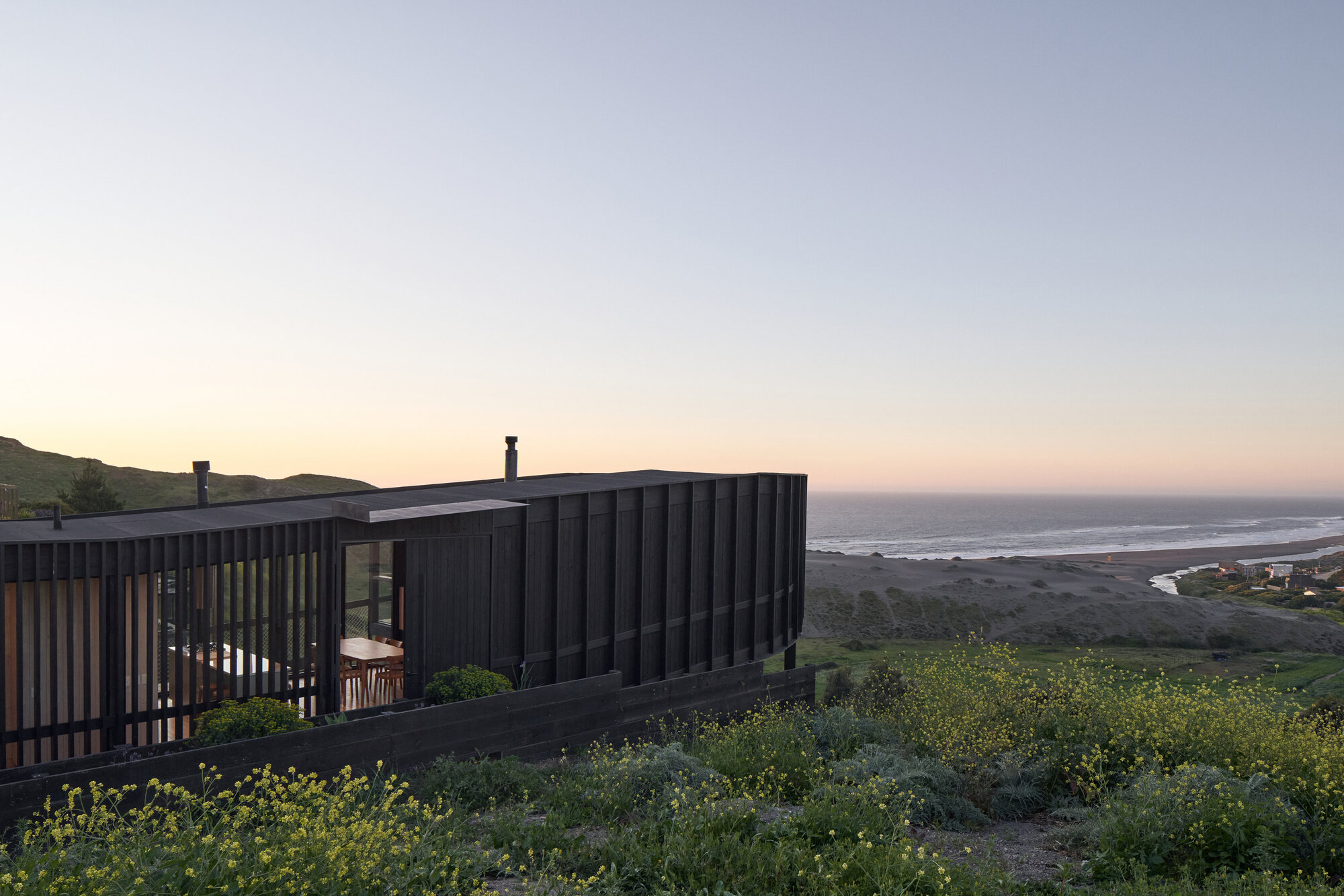 Slatted black timber facade with an open dining area, framed by yellow wildflowers and a pink dusk sky over the beach
