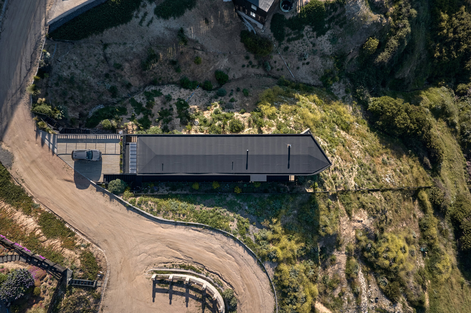 Drone view of the dark pitched roof on a steep slope, with a parked car beside a curving dirt road