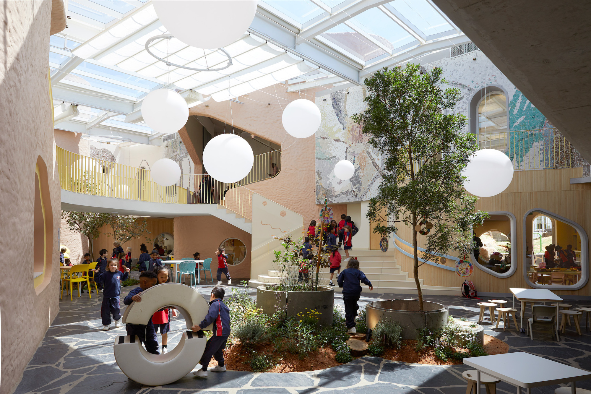 Daylit atrium with a potted tree, hanging white globe lights and children playing on a flagstone floor beneath a glazed roof