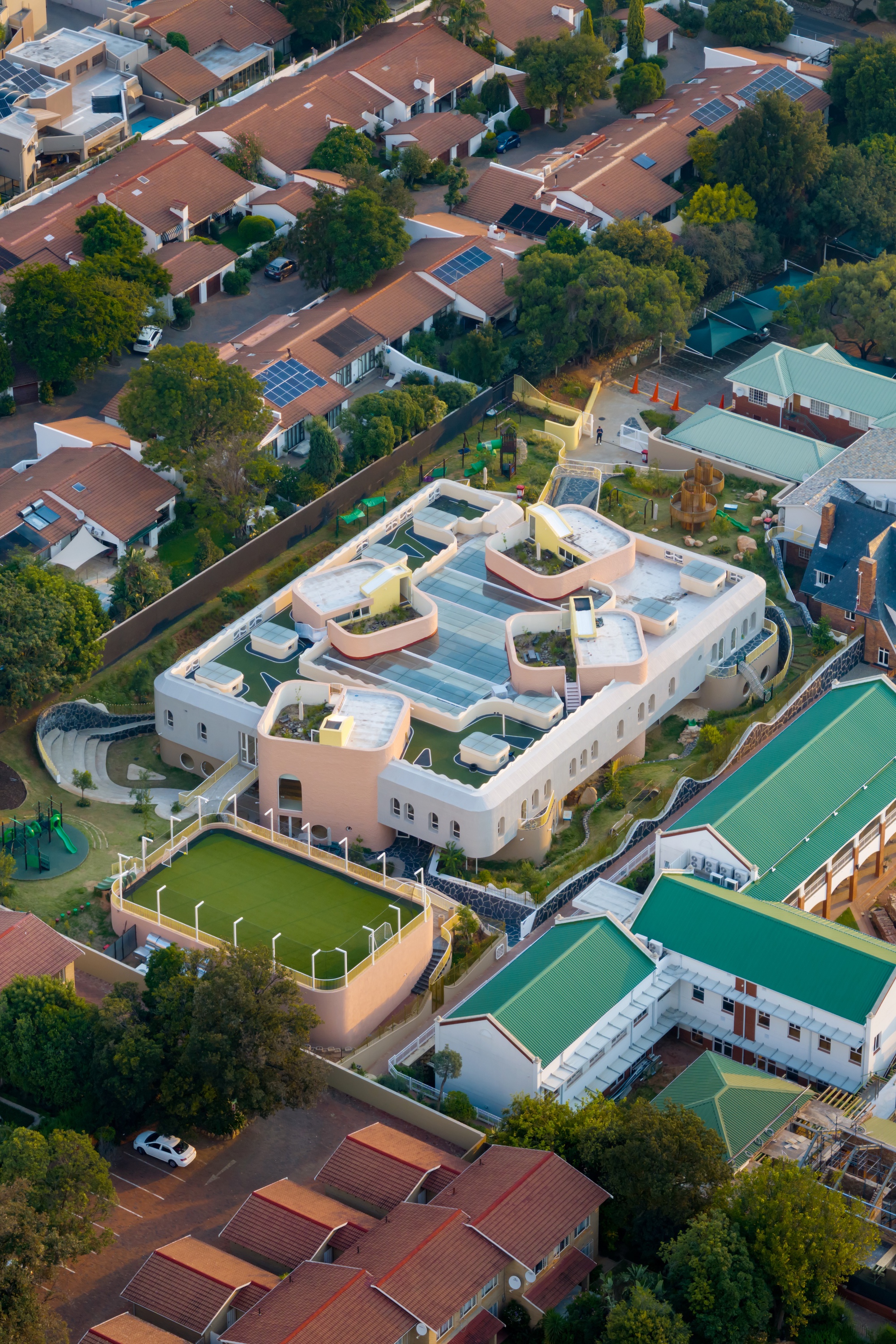Aerial view of the curved, pale rooftop terraces with planted beds, a rooftop play pitch and surrounding tiled-roof houses