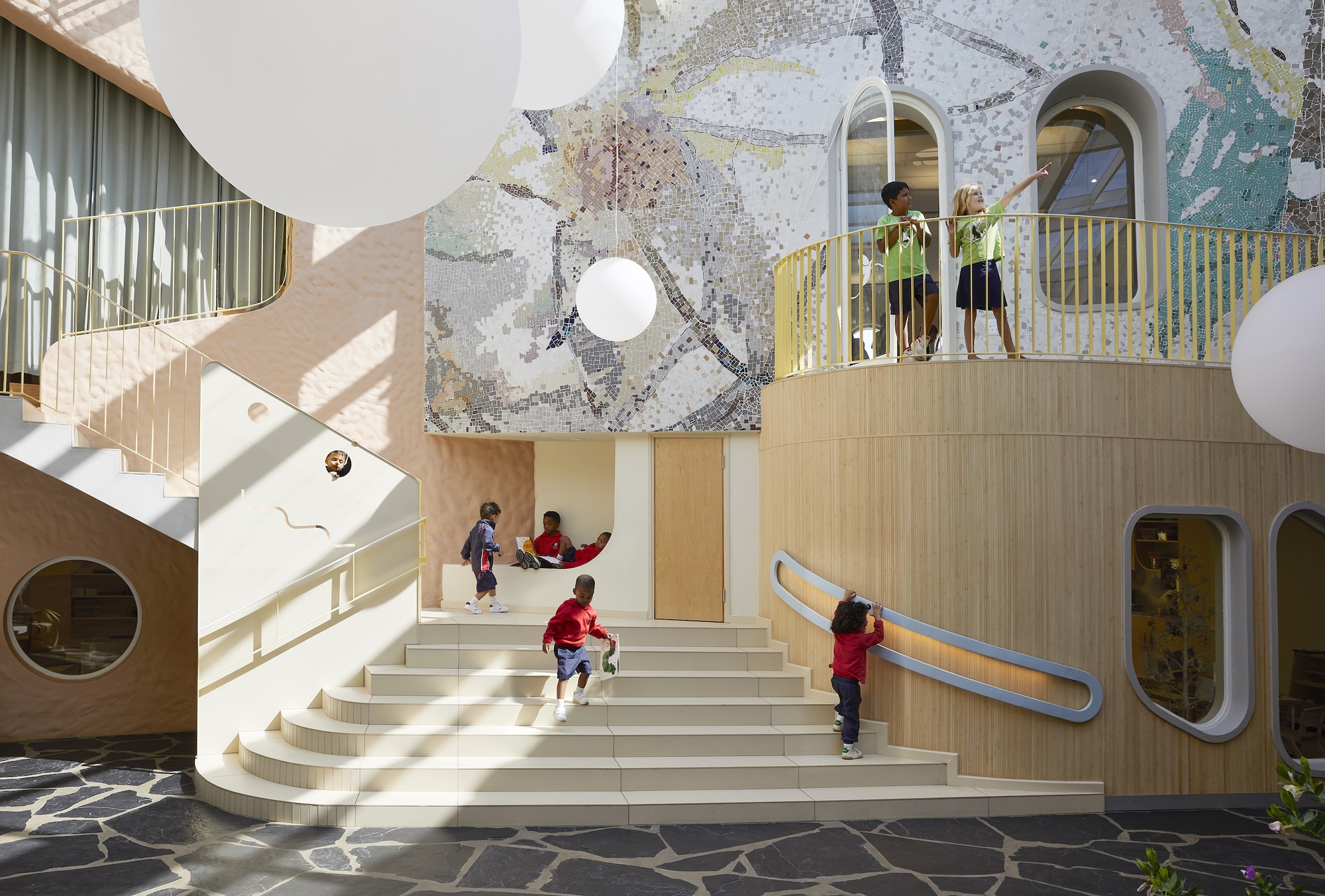 Wide stepped seating beside a timber-clad curved balcony and mosaic wall, with children climbing and reading in afternoon light