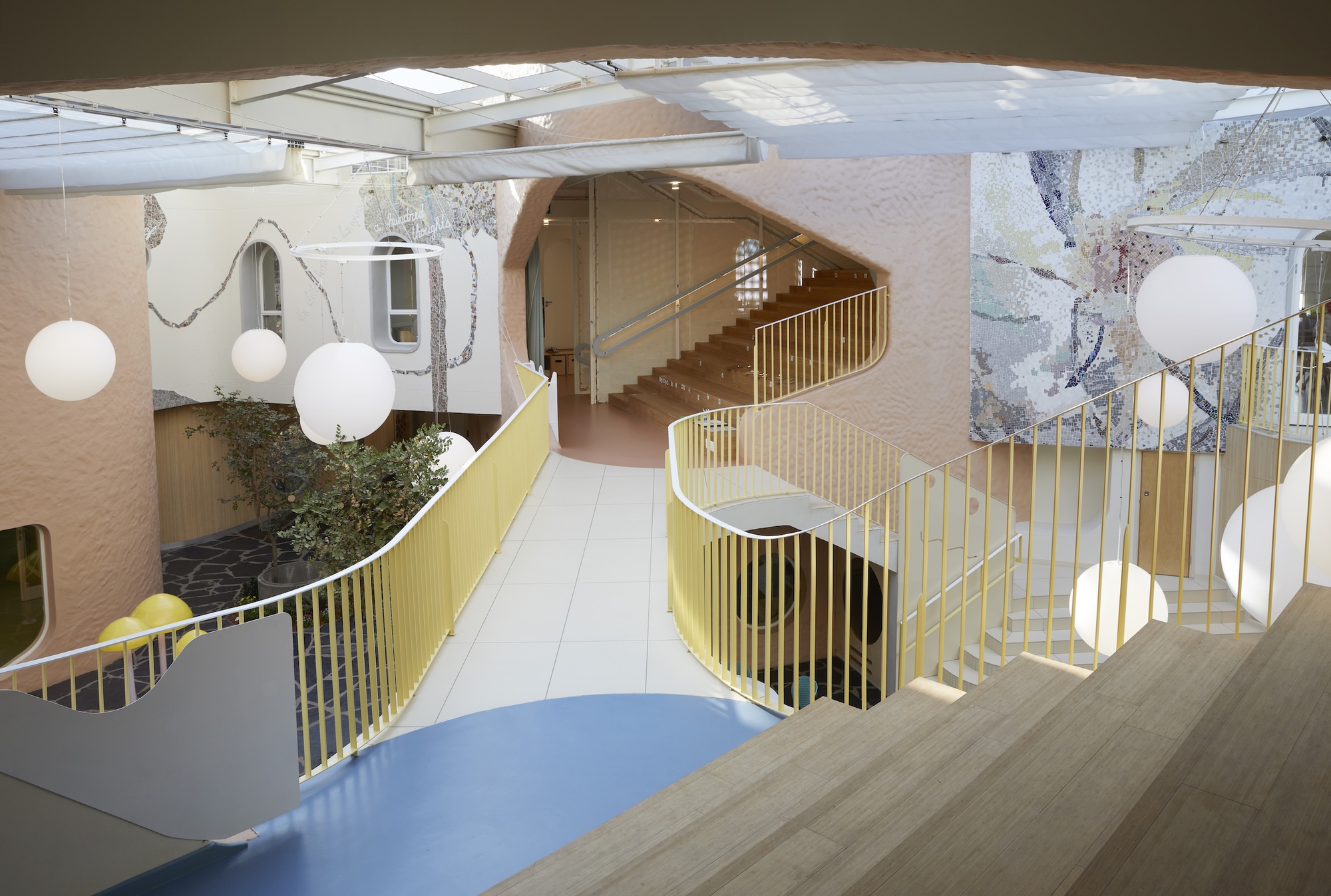 View down from a timber stair to a yellow-railed curving walkway, globe lights and a mosaic wall under a skylight