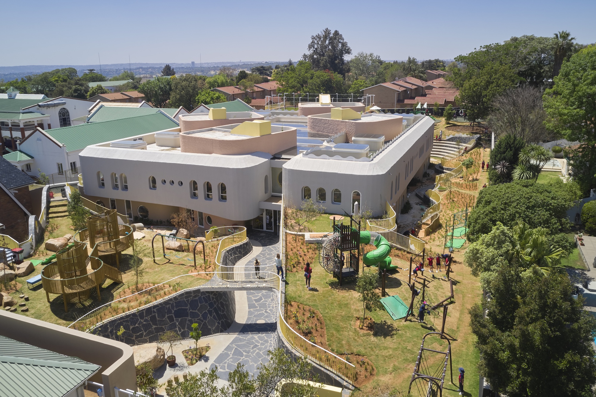Elevated view of the curved white facade with arched windows above a terraced playground, slides and flagstone path on a clear day