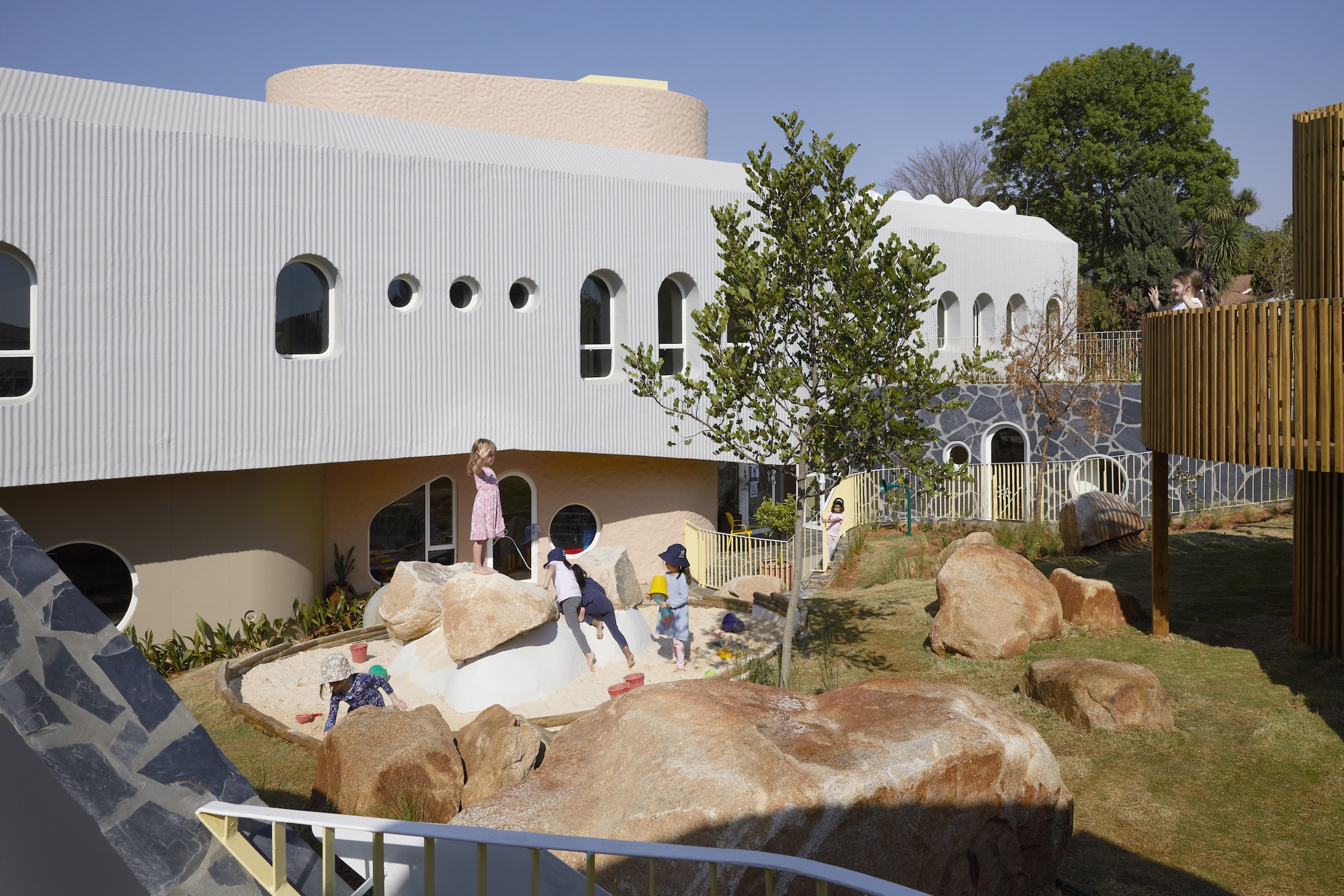 Corrugated white facade with round and arched windows above a sand pit and boulders where children play under a blue sky
