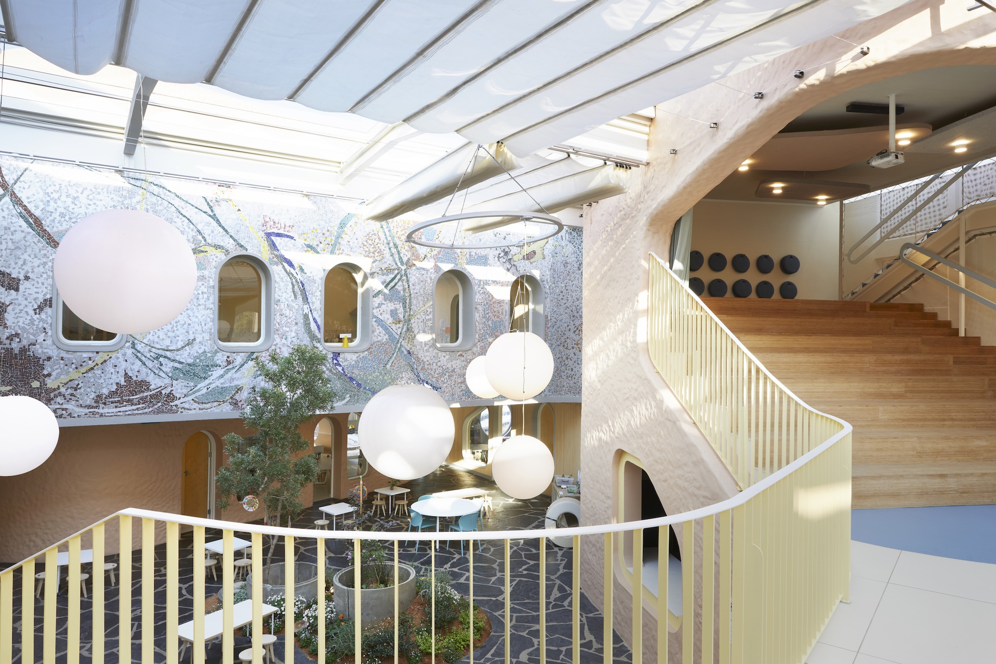 Atrium seen from a curved balcony, with pale yellow balusters, globe pendants and a mosaic wall under a fabric canopy