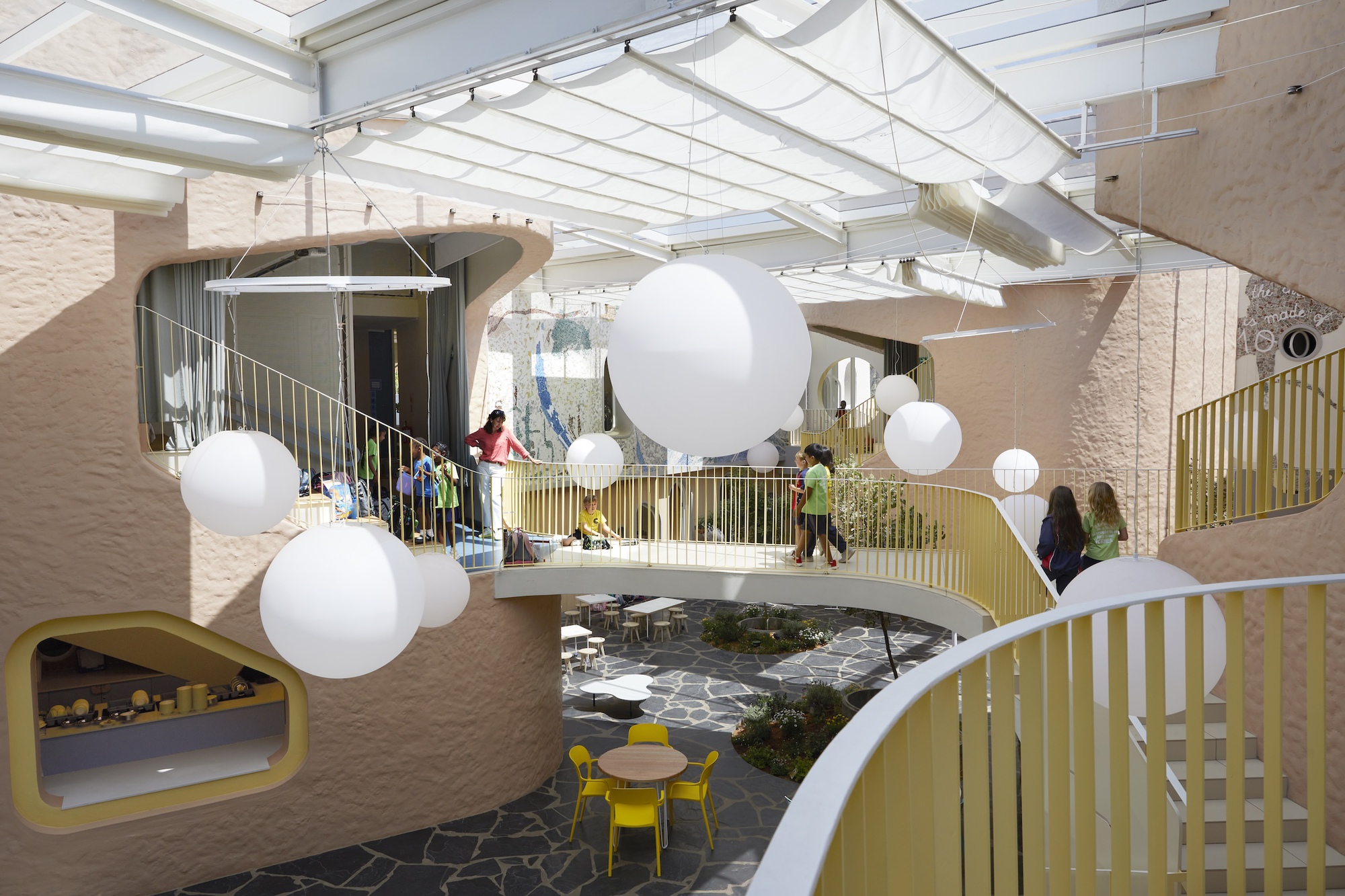 Curving bridge and yellow railings above a paved courtyard with round pendant lamps and children walking in daylight