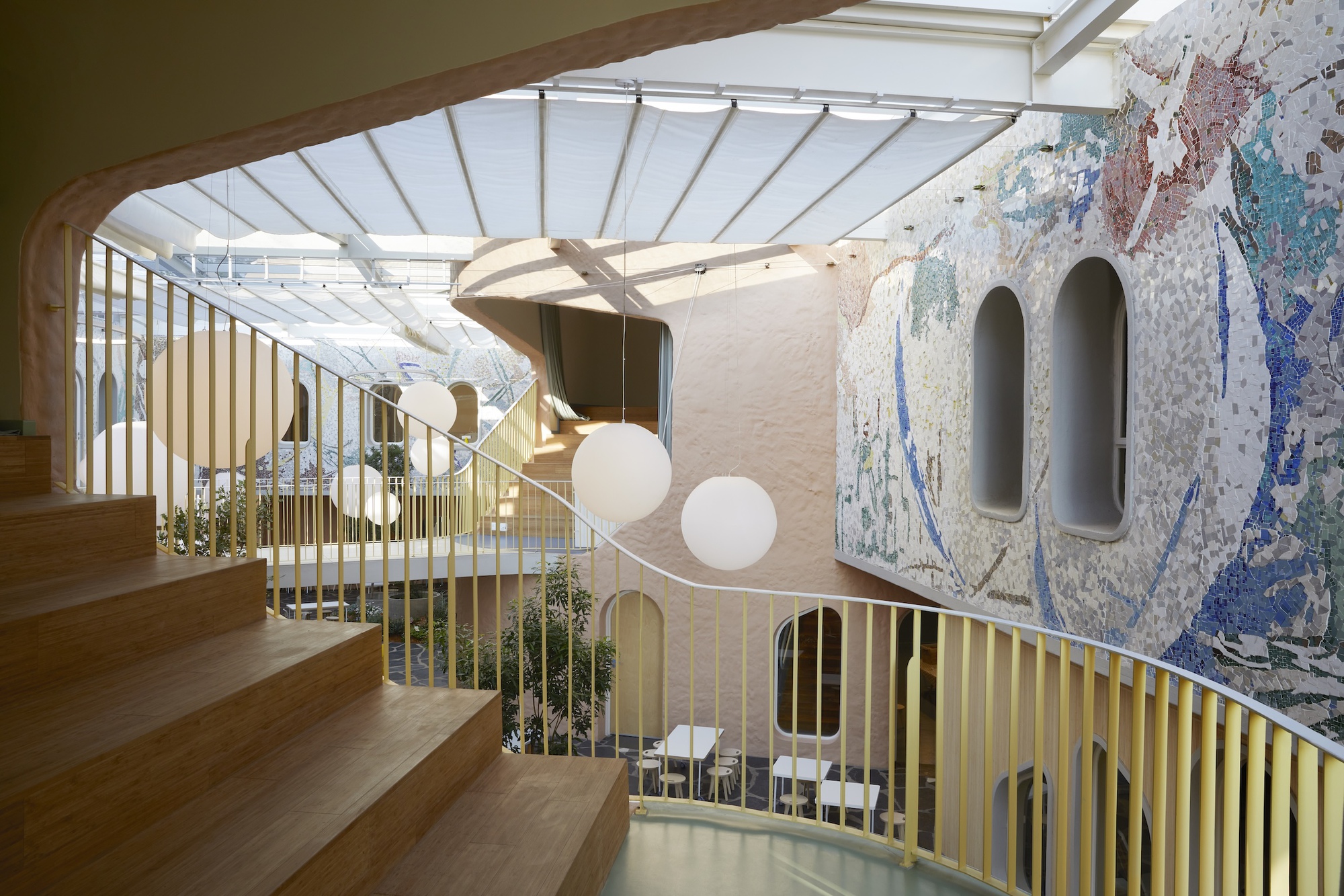 Timber steps beside a curved yellow balustrade, facing a mosaic wall with arched window niches under a sunlit canopy
