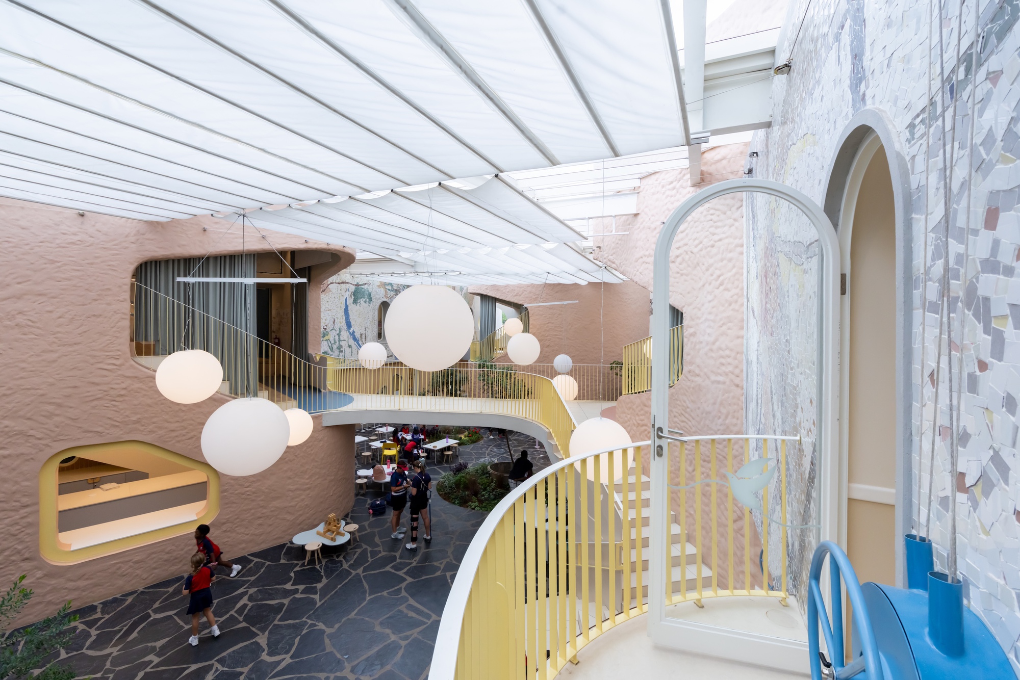Schoolchildren crossing a stone-paved atrium floor beneath white globe lights, with a glazed arched door and mosaic wall at right