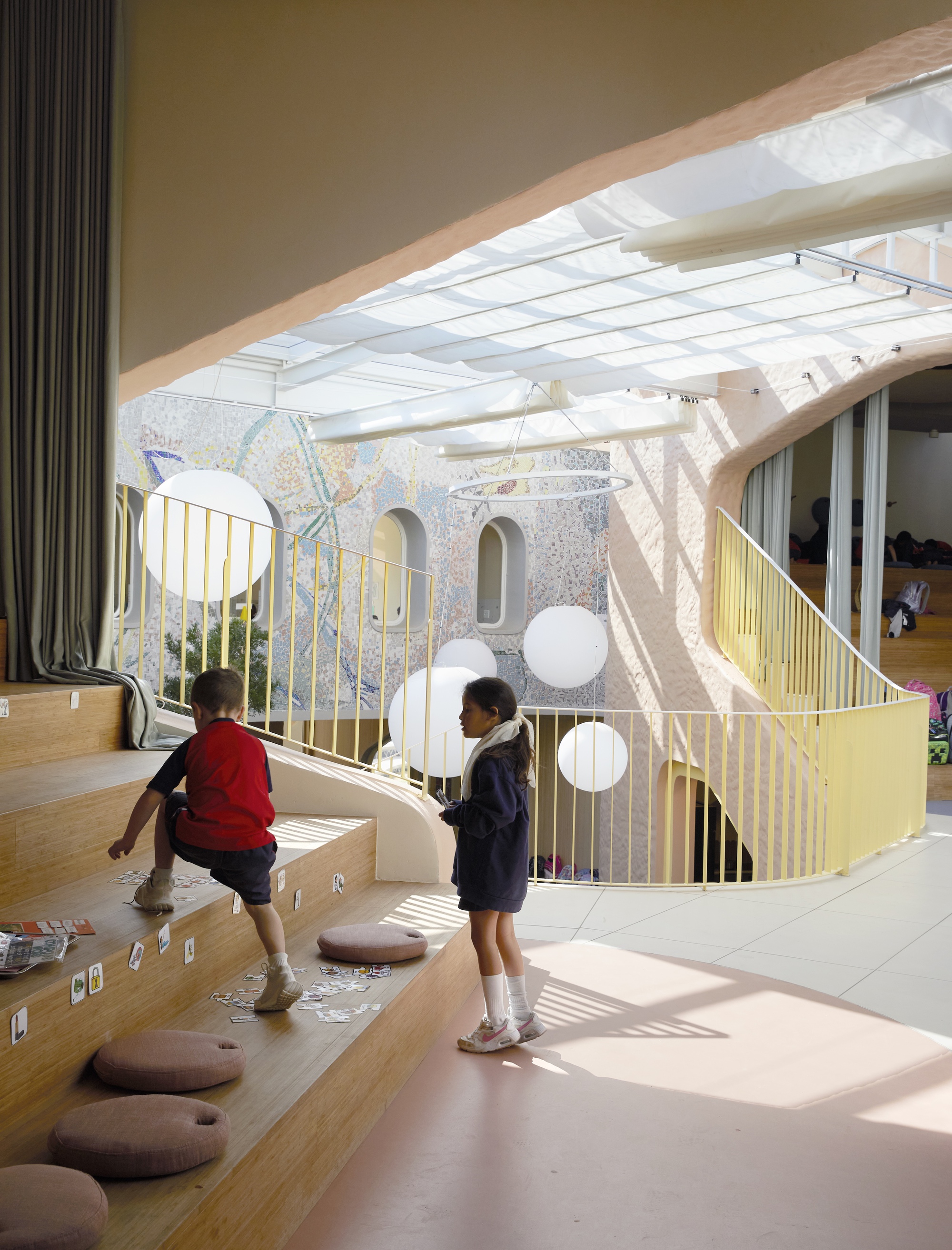 Two children on timber tiered steps with round floor cushions, with the atrium's mosaic wall and globe lights behind in afternoon sun