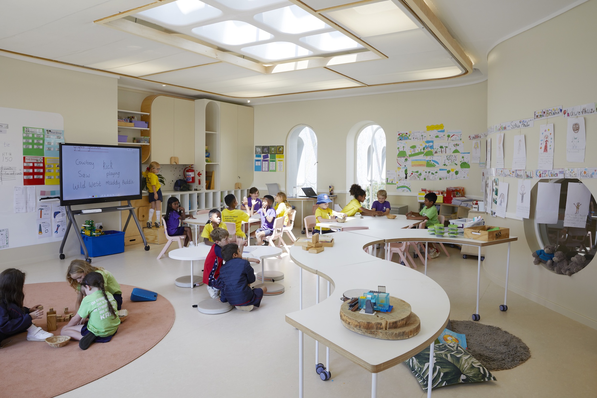 Classroom with a curving white table, children at work, arched windows and a skylight cluster in the ceiling