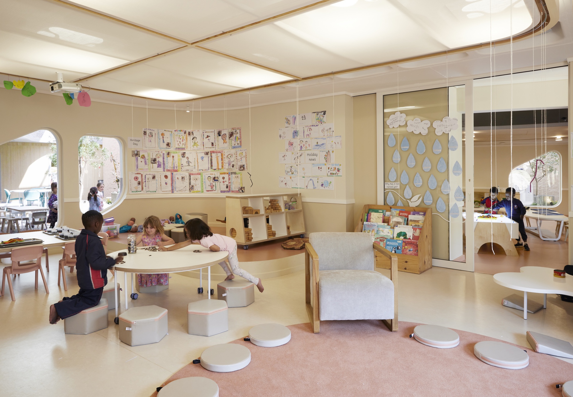 Classroom with round pink rug, hexagonal stools, children's drawings on the wall and arched openings, lit by soft ceiling panels