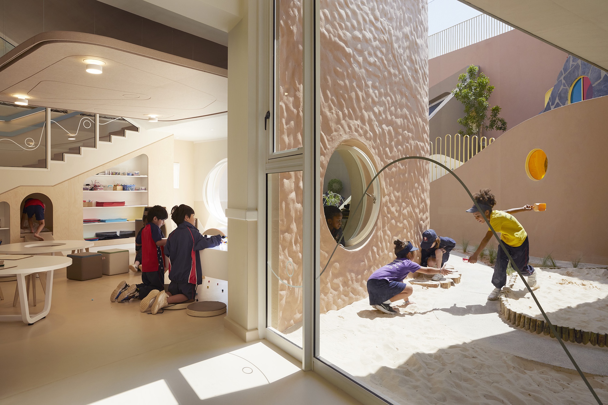 Children playing in a sand courtyard beside a textured pink curved wall with a round porthole window, seen through glass in bright sun