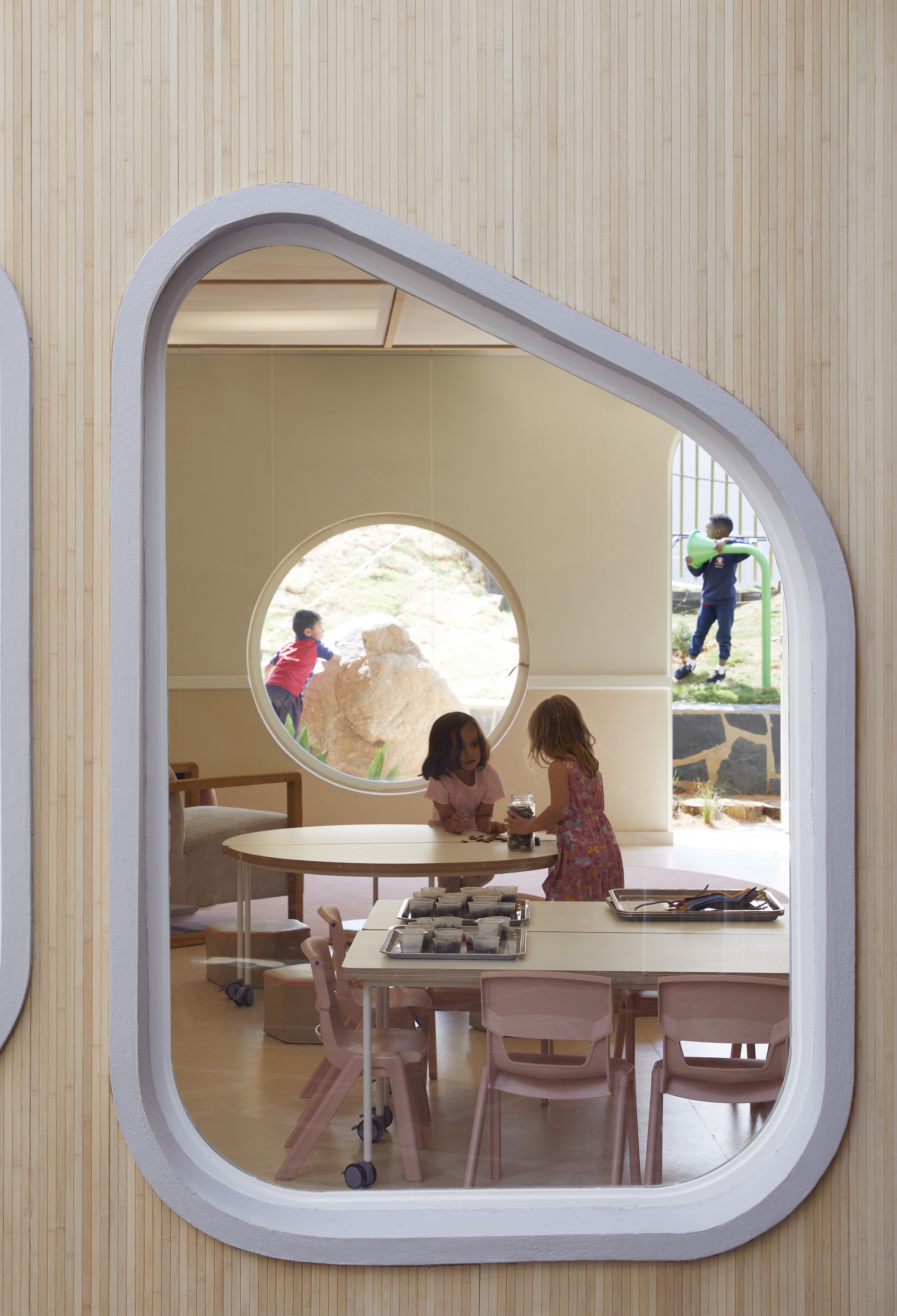 Rounded window cut into a timber slat wall, framing two girls at a table and a child climbing a boulder beyond
