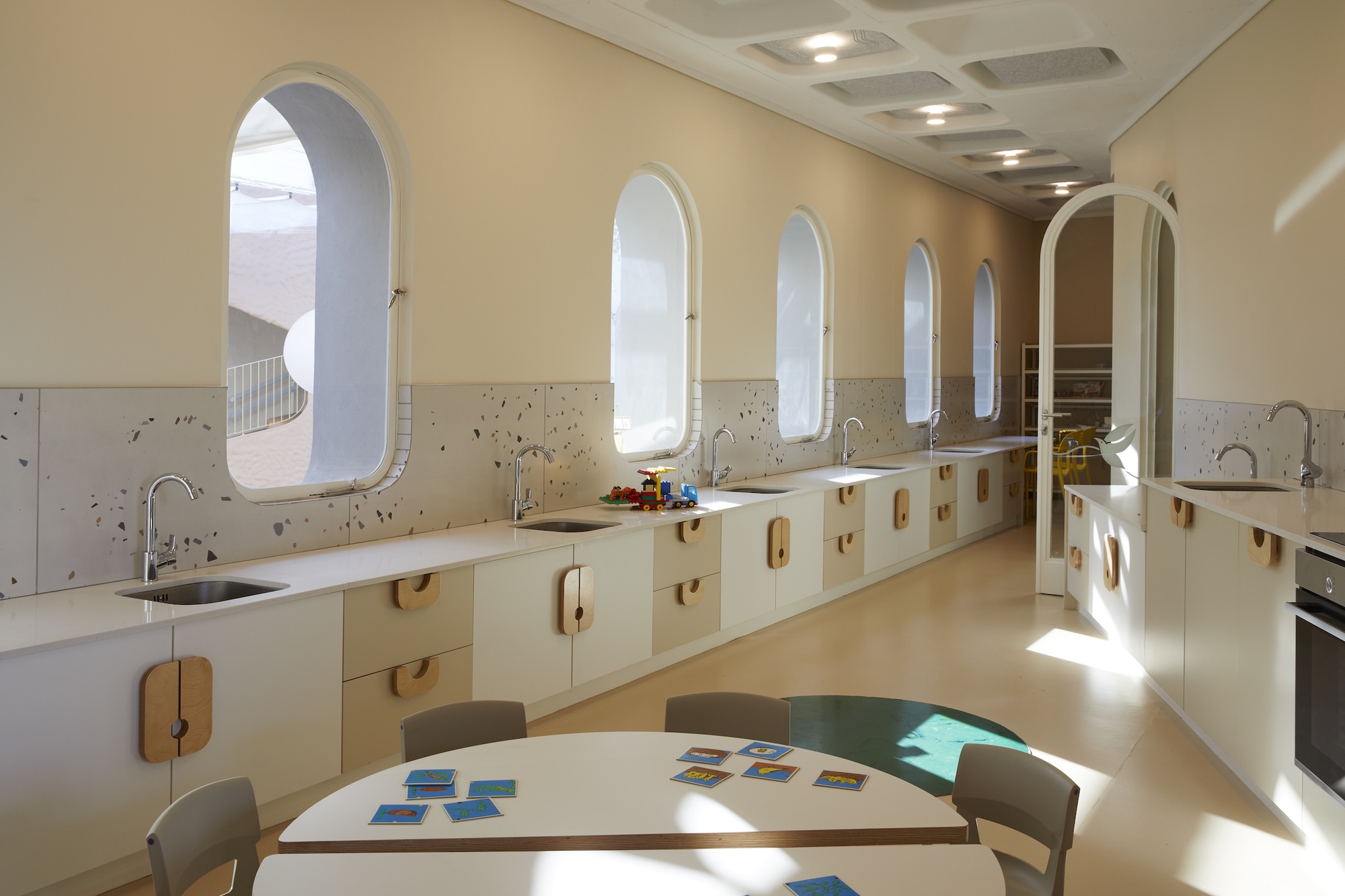 Long kitchen counter with sinks and arched windows under a coffered ceiling, with a round table set with picture cards in the foreground