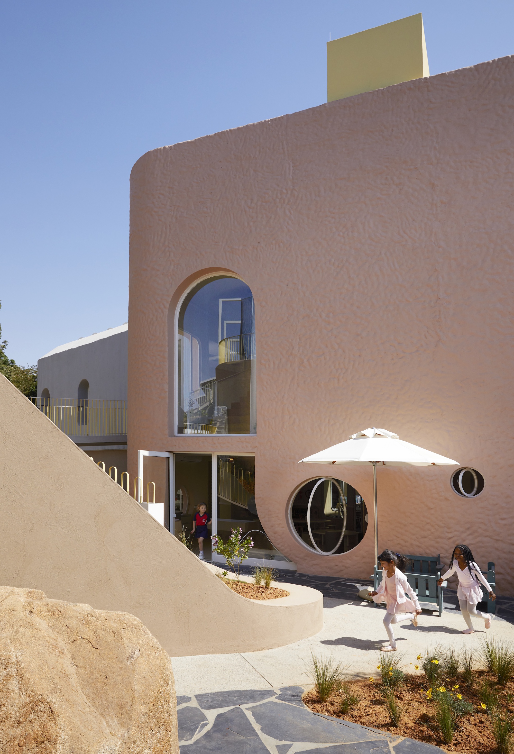 Curved pink textured facade with an arched window and round portholes, as two girls run past a white umbrella under clear blue sky