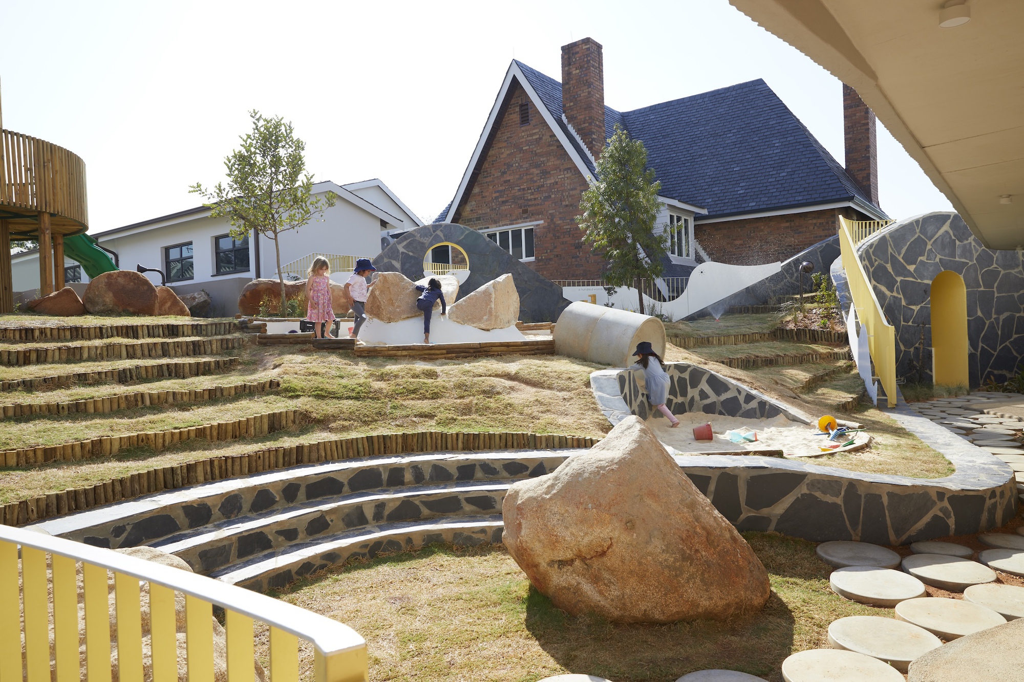 Terraced grass amphitheatre with log edging, stone-clad walls, boulders and yellow railings, children playing in front of a brick house in afternoon sun