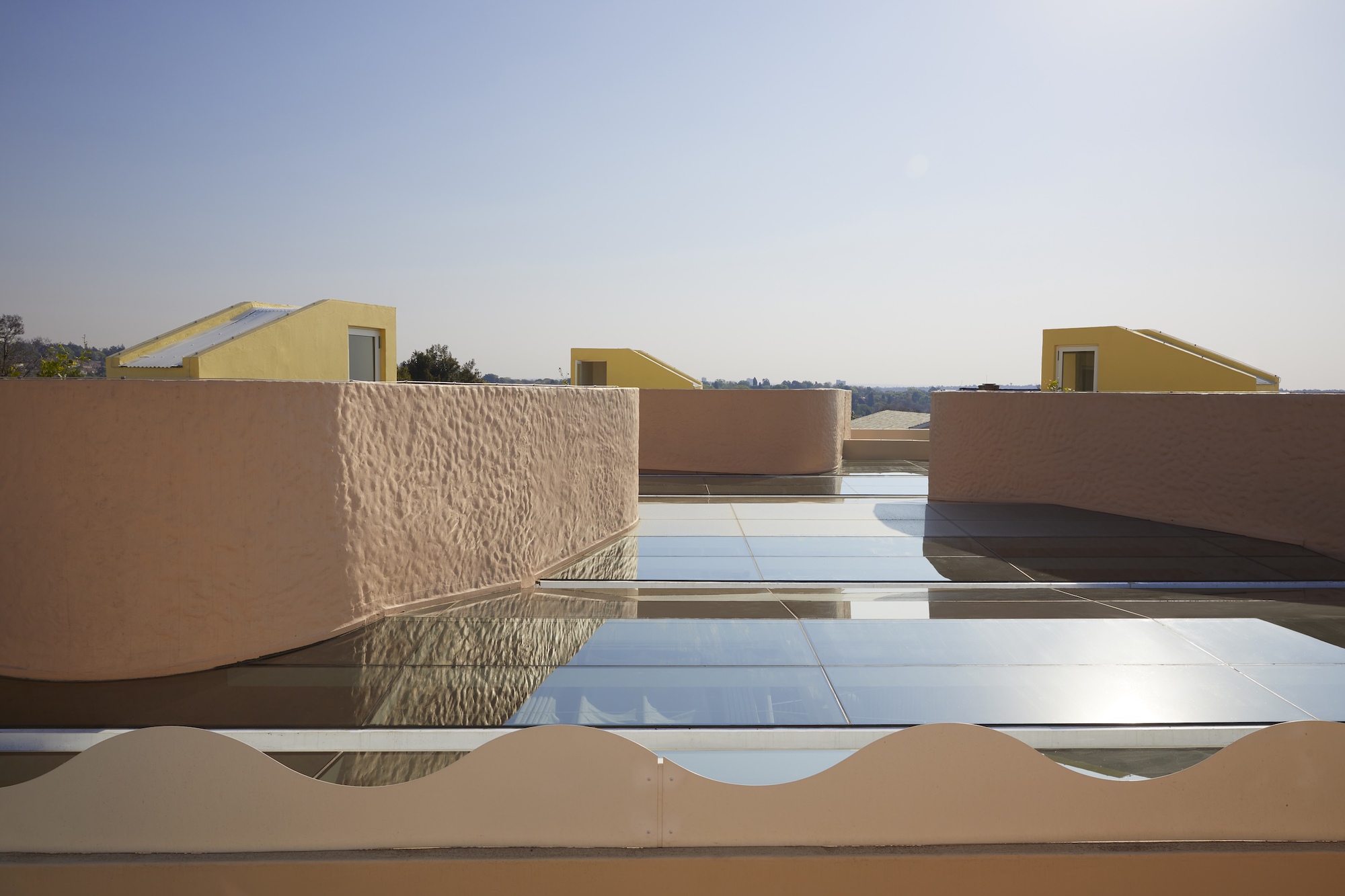 Rooftop glass panels between rounded pink textured parapets, with yellow dormer volumes on the horizon in clear afternoon light
