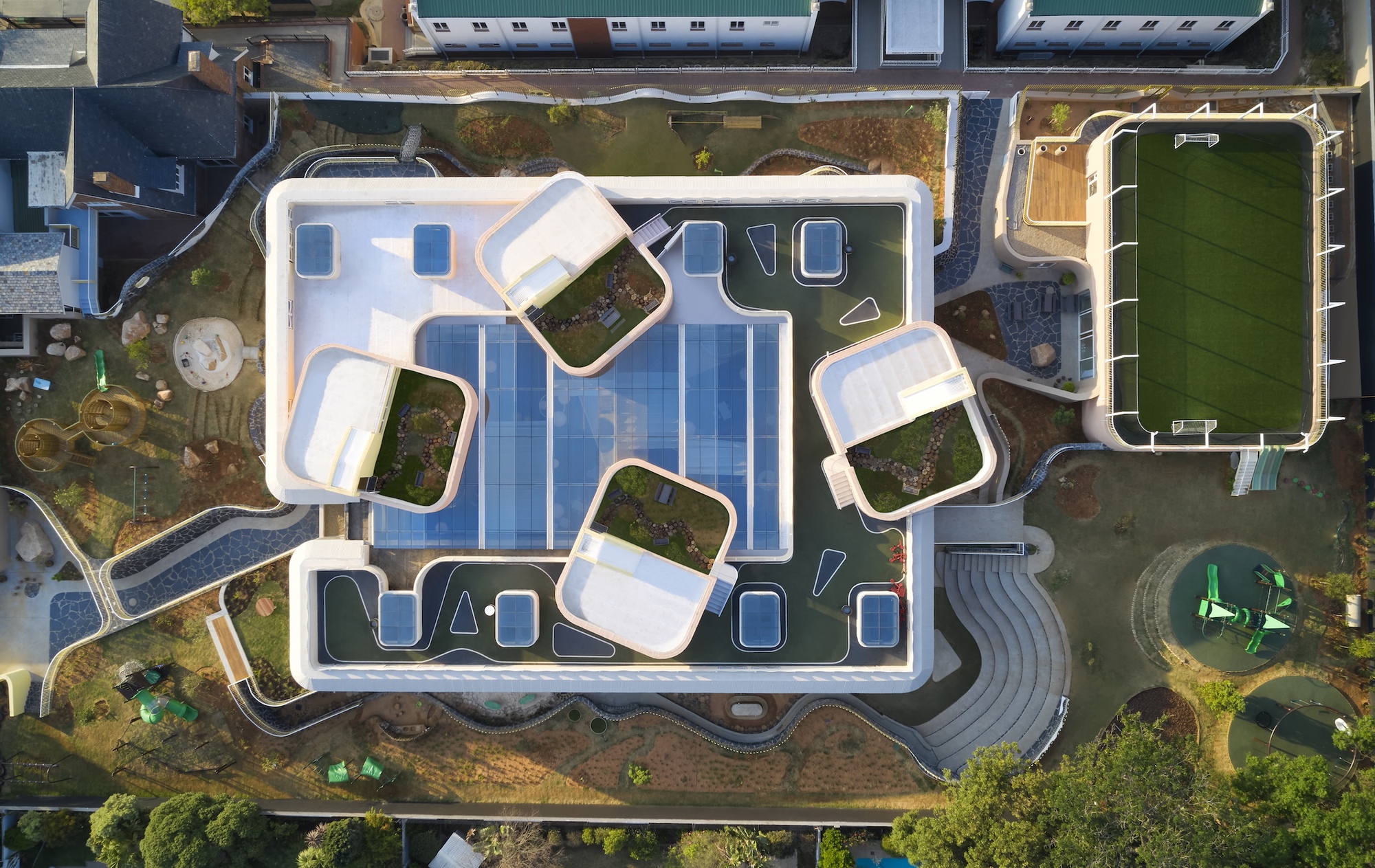 Aerial view of the roof with four rounded planted pavilions around a central skylight, beside a green sports pitch in low sun