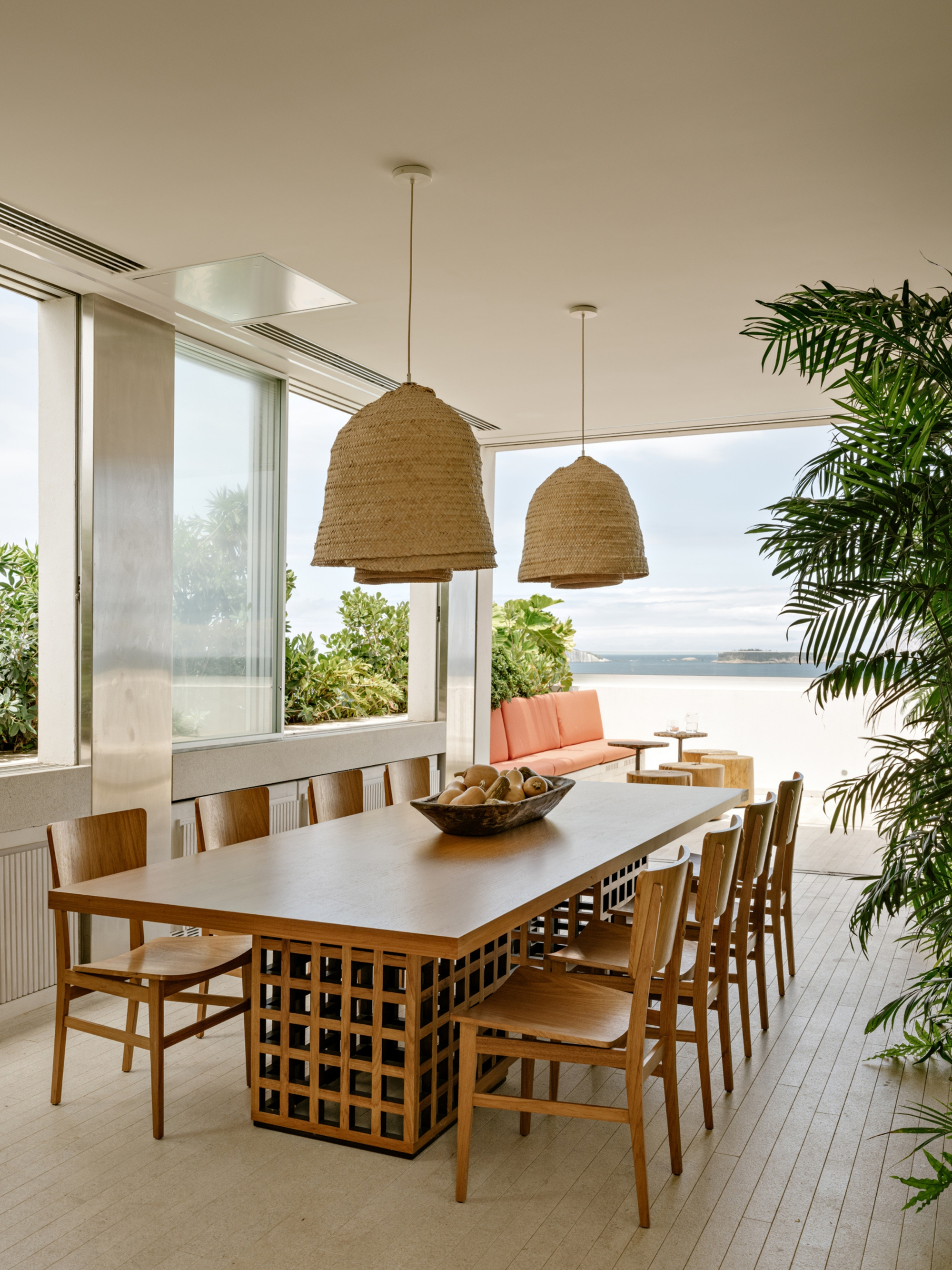 Long timber dining table with a lattice base under two woven pendant lamps, opening toward a beach and palm fronds