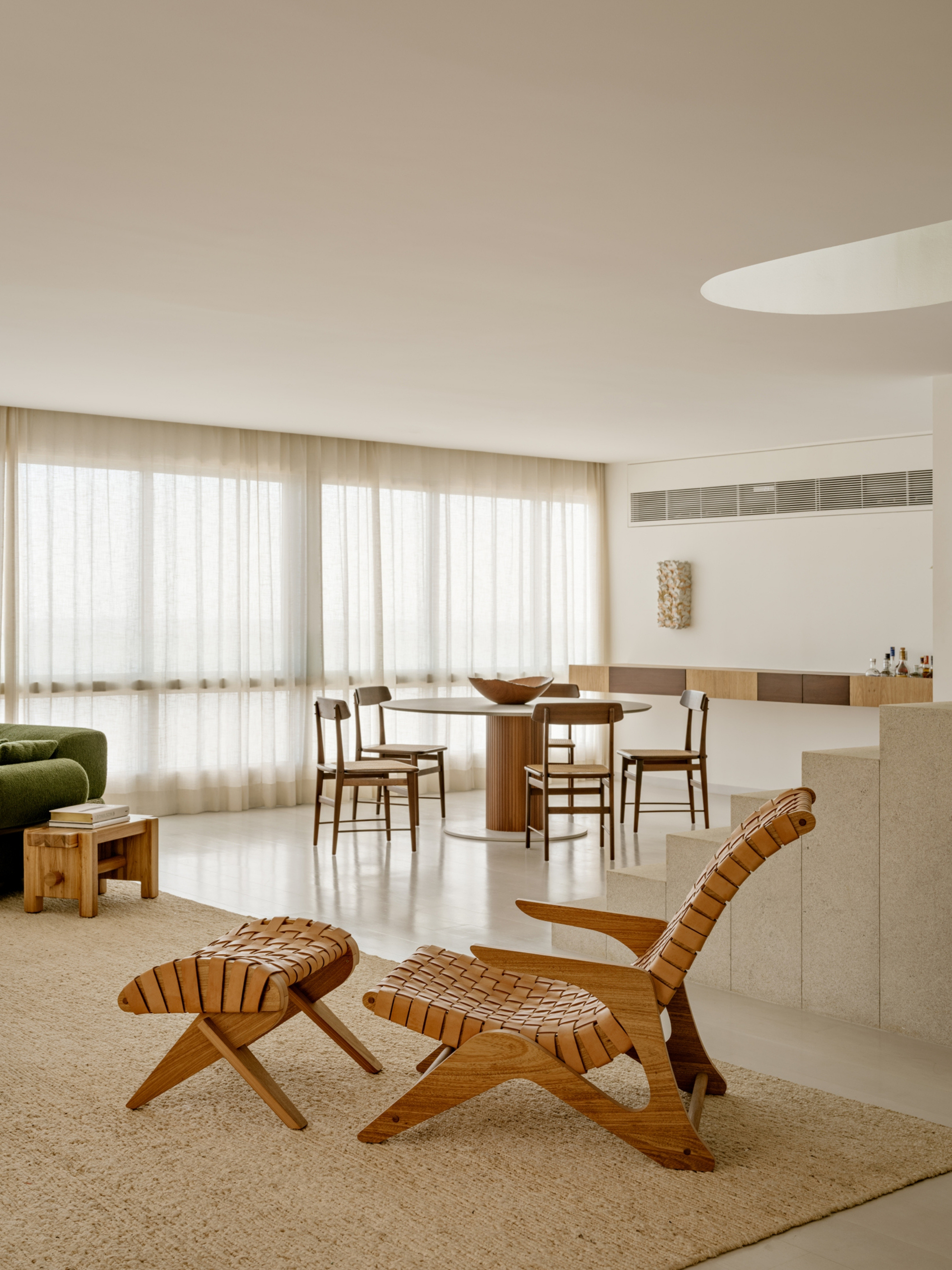 Woven leather lounge chair and ottoman on a jute rug, with a round dining table beside sheer curtains in the background