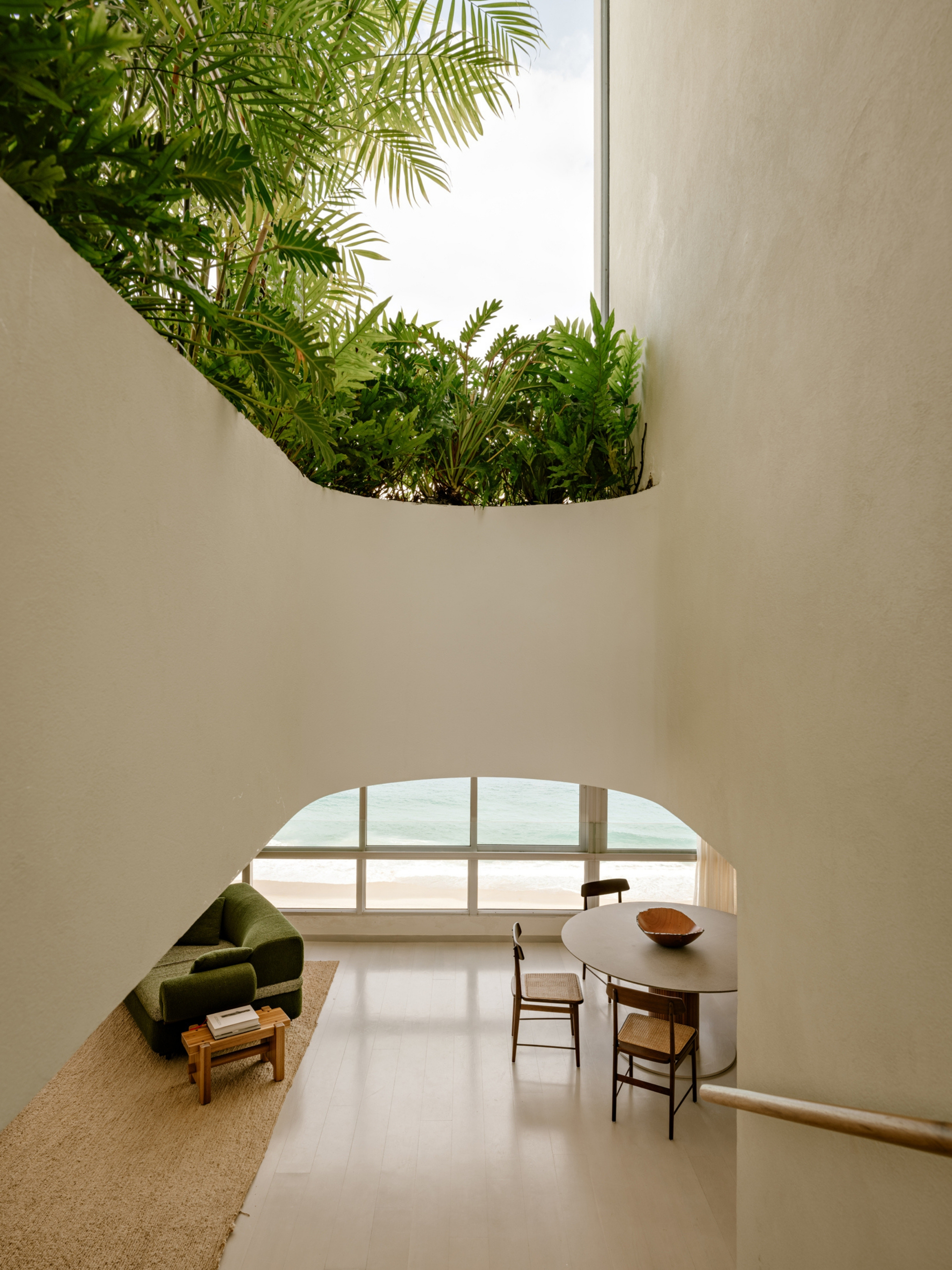 Curved plaster wall framing a planted terrace above an arched window, with the sea visible beyond a green sofa and dining table
