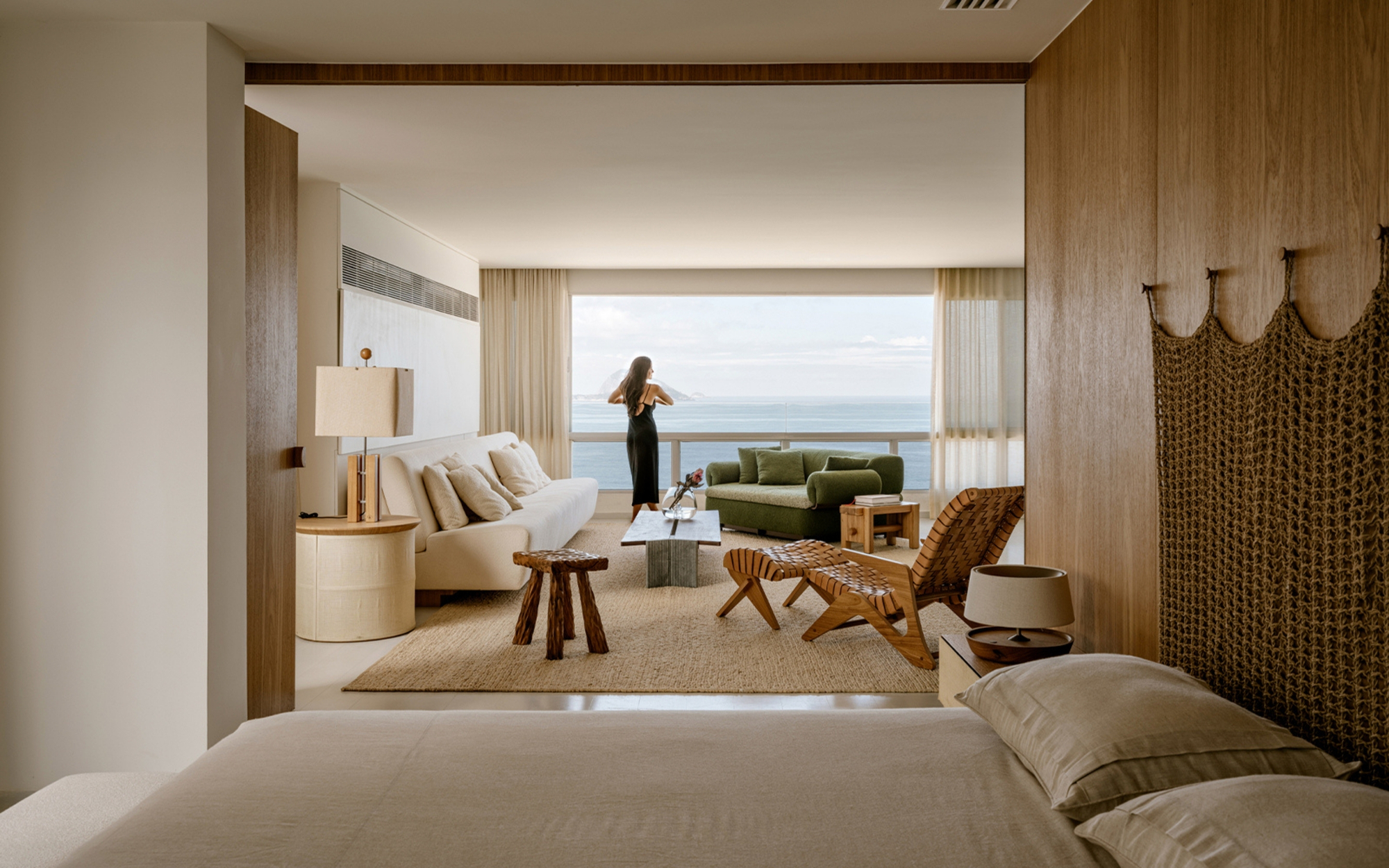 Bedroom looking through to a living area, where a woman in a black dress stands at sheer curtains facing the ocean