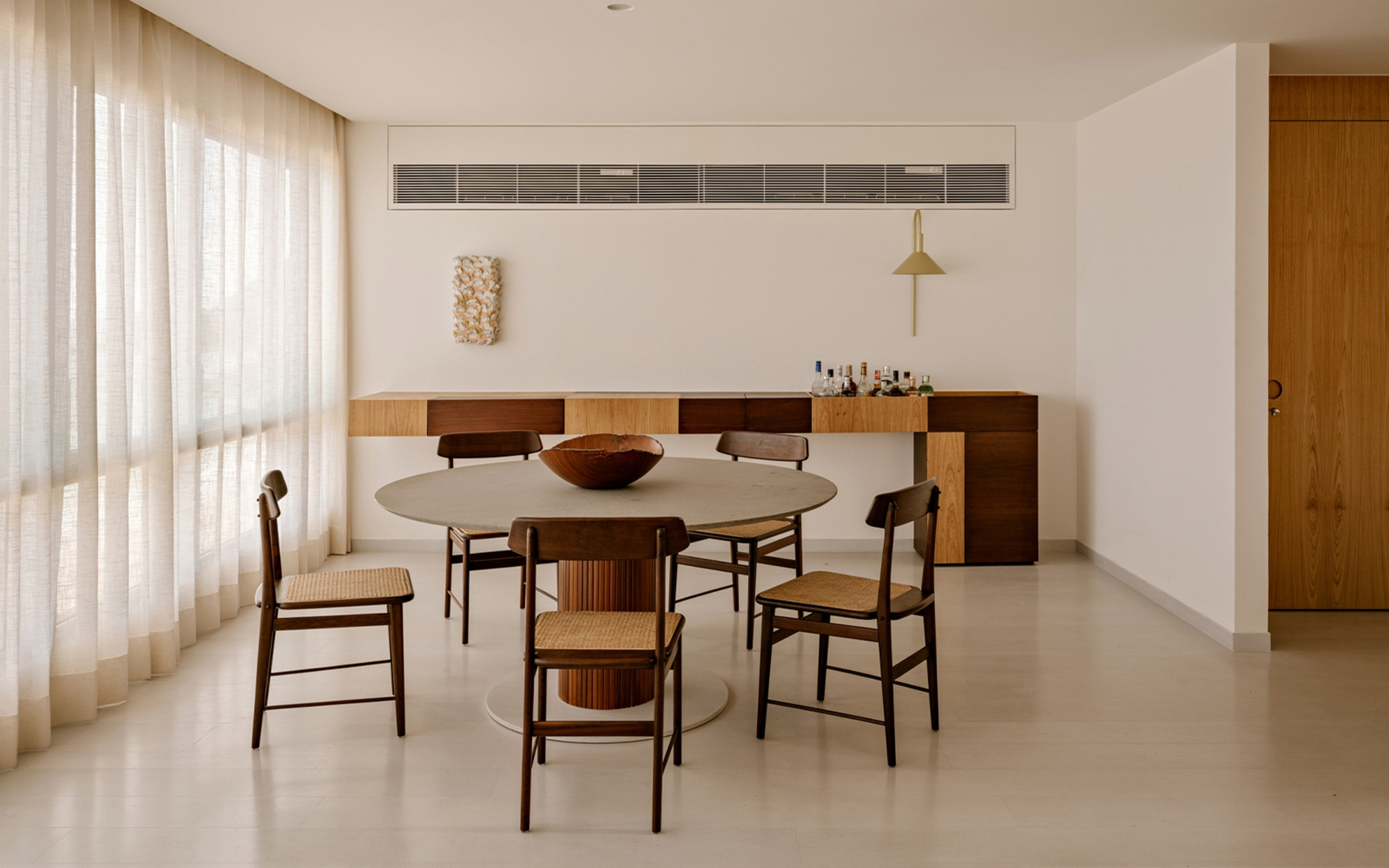 Dining area with a round stone-top table, caned wooden chairs and a timber sideboard beside floor-to-ceiling sheer curtains