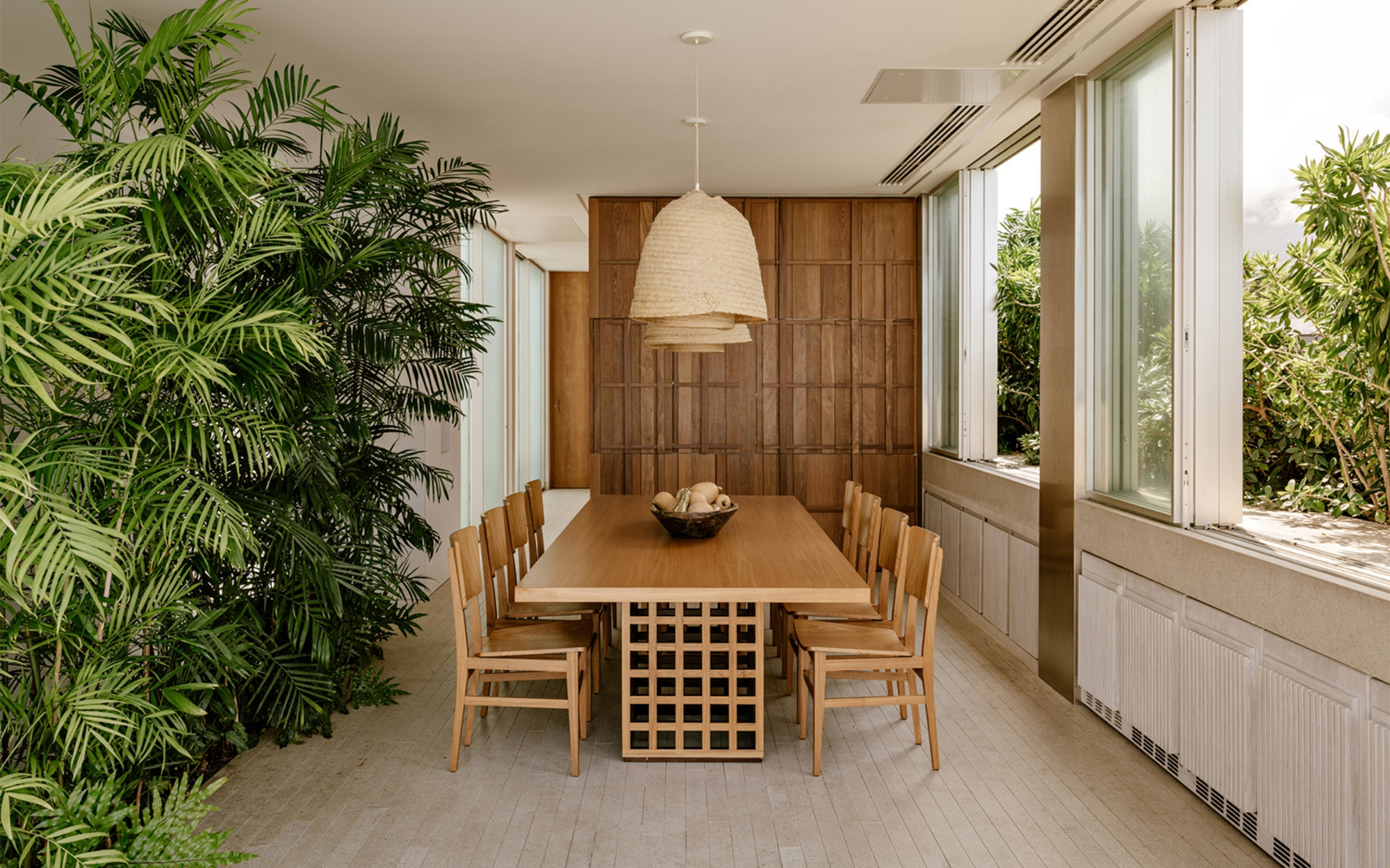 Long timber dining table under a woven pendant lamp, with tall palms on the left and sliding windows opening to foliage